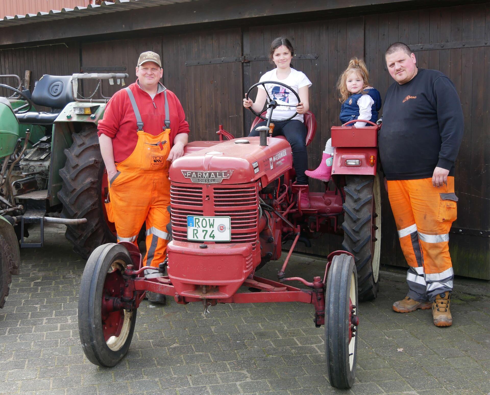 Zwei Kindern sitzen auf einem kleinen roten Traktor. Daneben stehen zwei Männer.