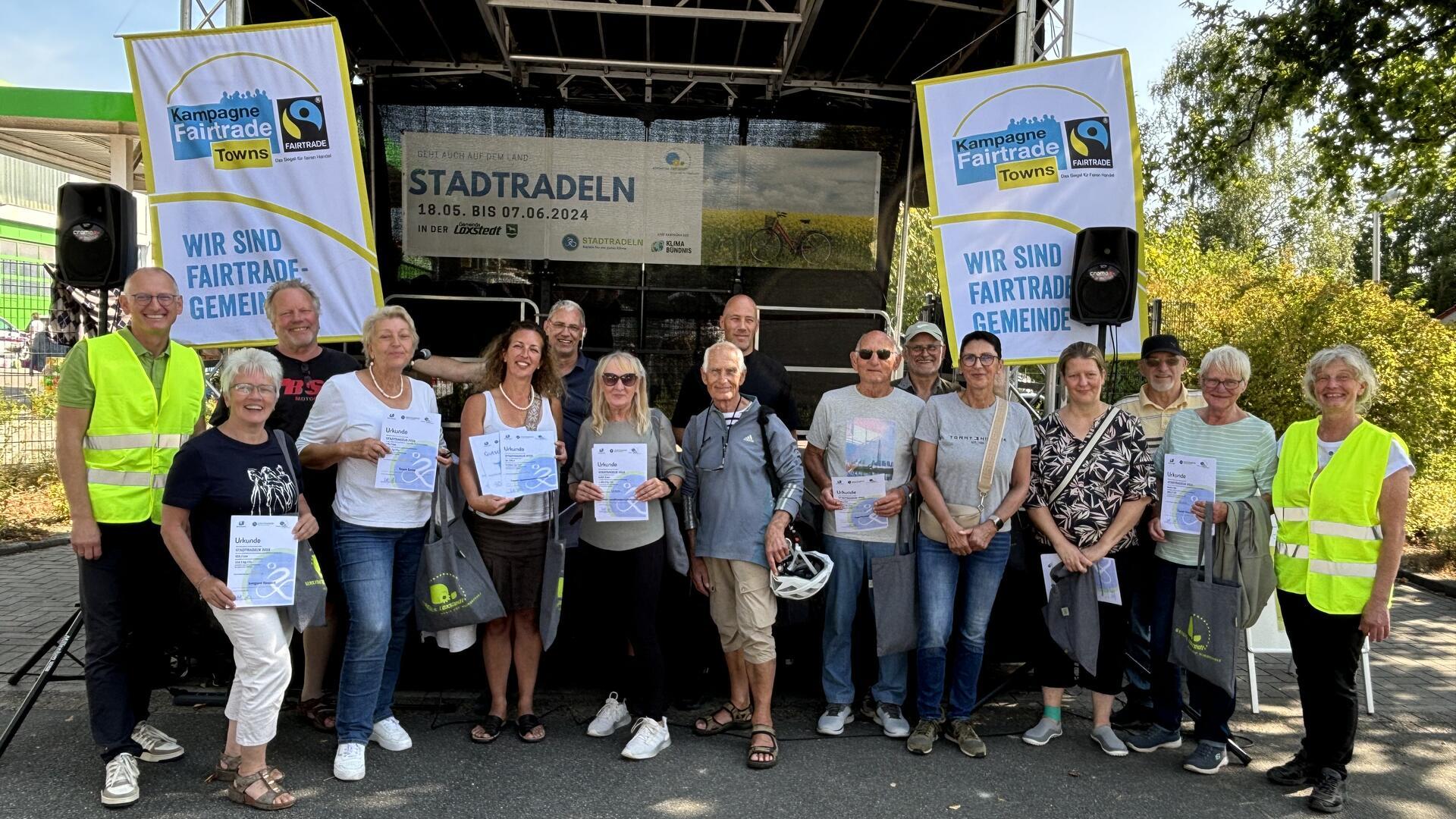 16 Frauen und Männer stehen nebeneinander vor einer Bühne. Einige halten Urkunden in Händen.