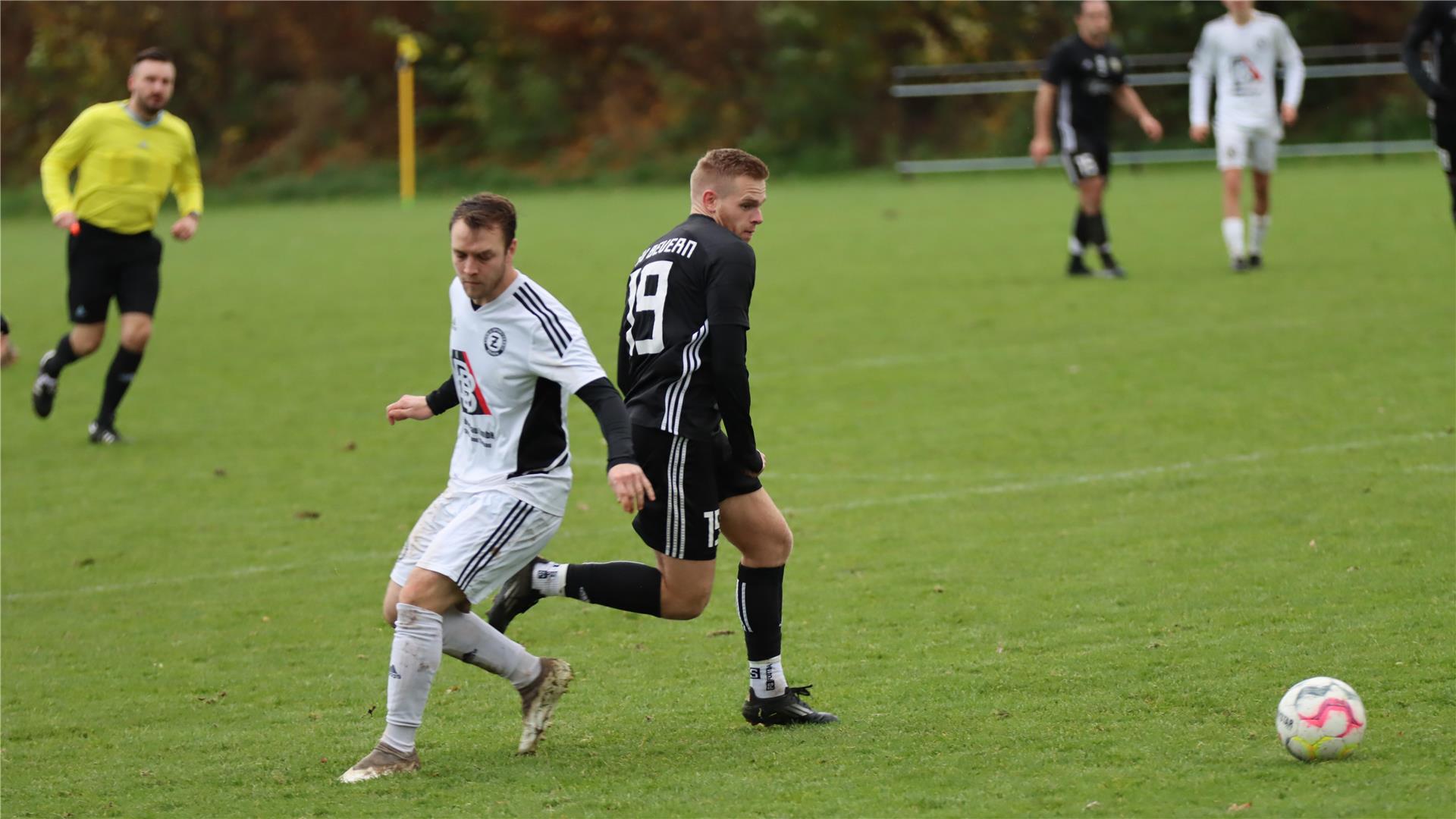 Zwei Männer in weißen und schwarzen Trikots laufen neben einem Ball.