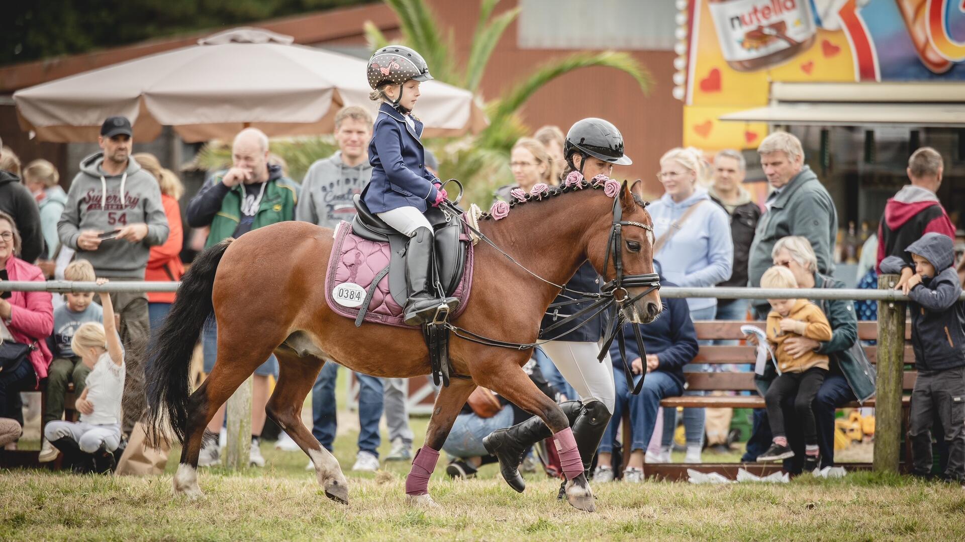 Ein kleines Mädchen auf einem Pony und Zuschauer im Hintergrund.