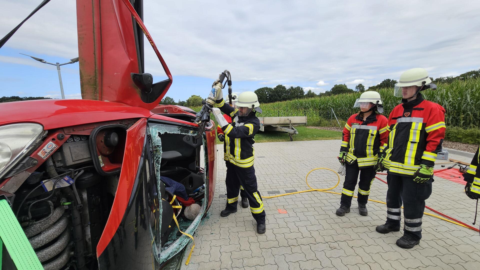 Ein Auto liegt auf der Seite. Ein Feuerwehrmann bearbeitet die Karosserie mit einem hydraulischen Rettungsgerät, während zwei Feuerwehrleute ihn dabei beobachten.