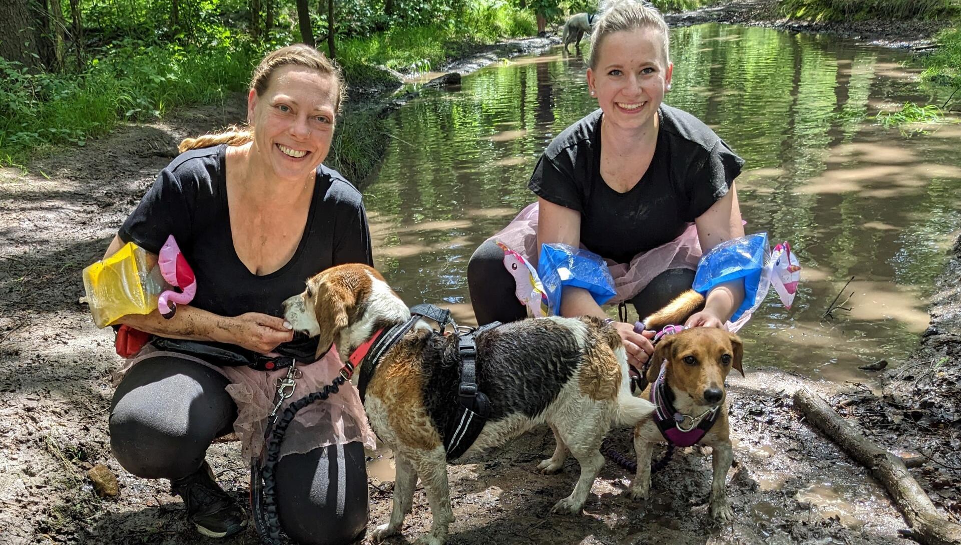 Zwei Frauen mit Schwimmflügeln und rosa Tutus knien mit ihren Hunden vor einem kleinen Teich