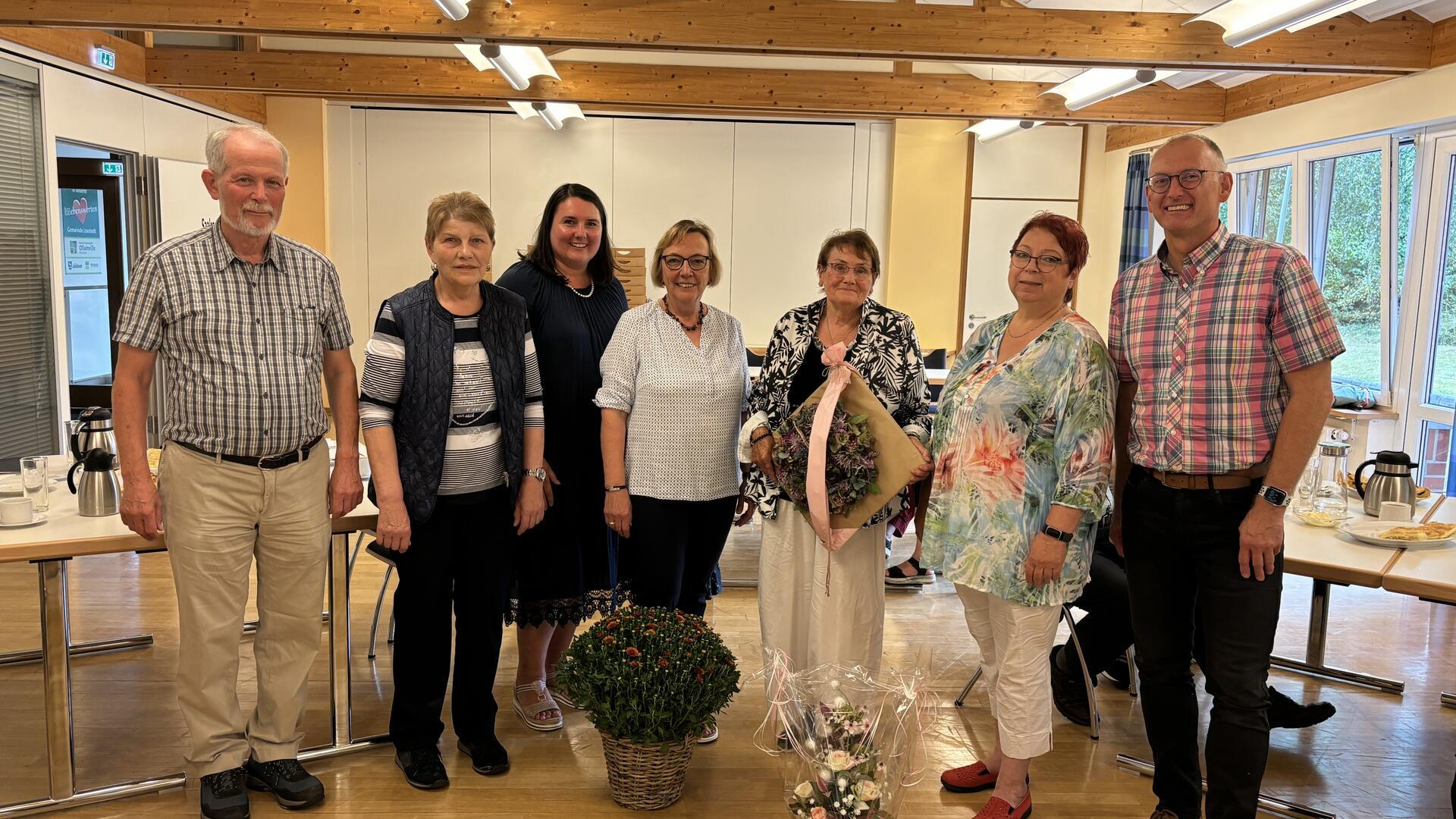 Fünf Frauen und zwei Männer stehen nebeneinander in einem Saal. Eine der Frauen hält ein Geschenk in Händen.