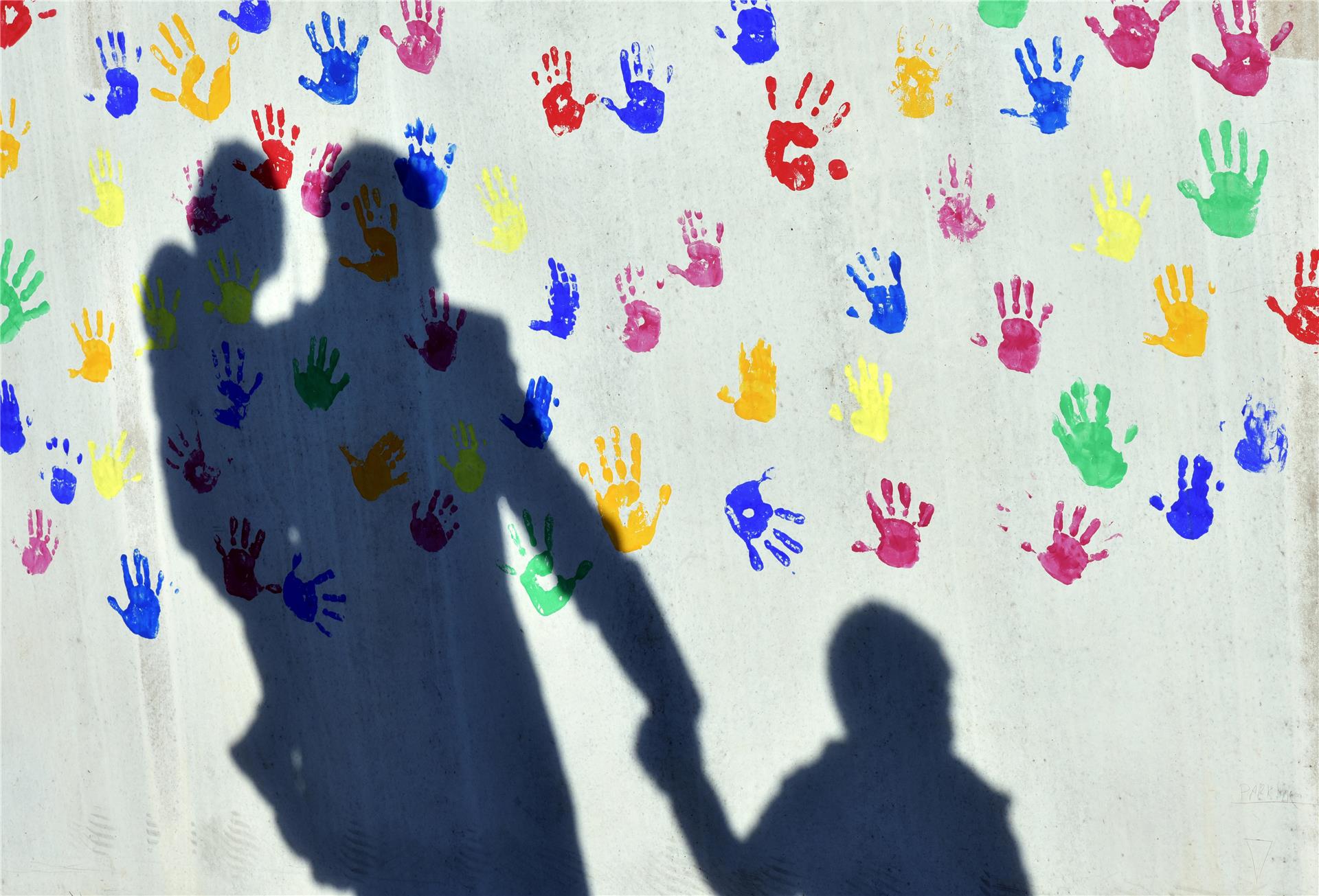 Ein Mann mit einem Kind auf dem Arm und einem an der Hand wirft einen Schatten auf eine mit bunten Handabdrücken bemalte Wand einer Kindertagesstätte