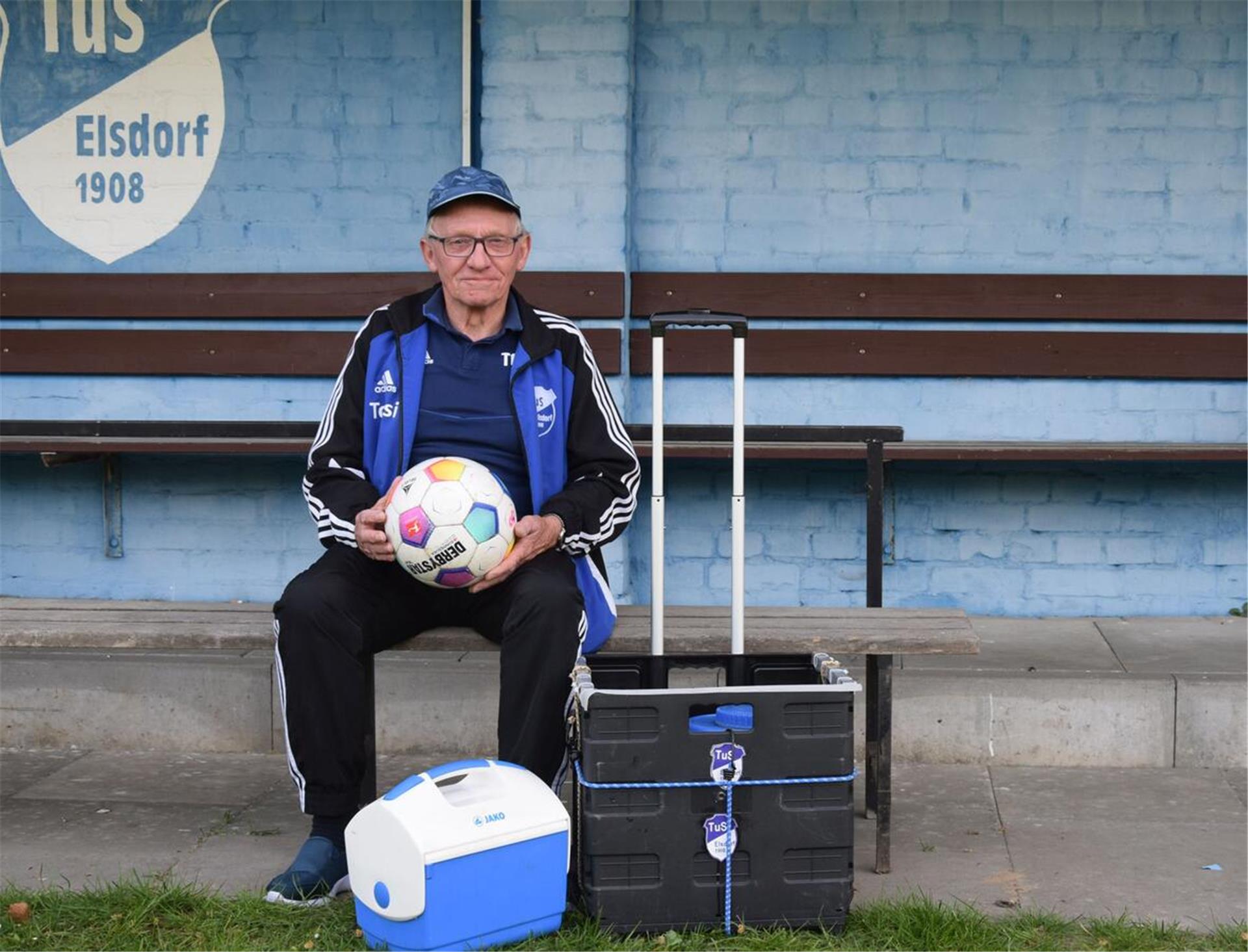 ein alter Mann in Trainingsanzug, mit einer Kappe und einer Brille, sitzt auf einer Bank und hält einen Ball in der Hand