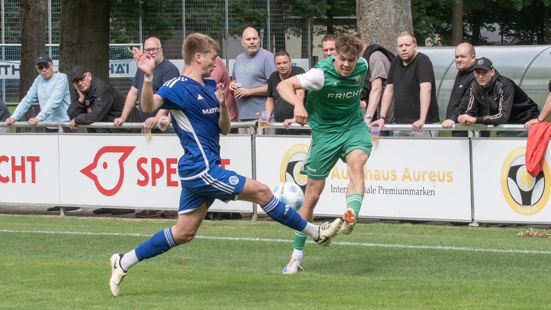 Zwei Männer kämpfen um einen Ball und mehrere Männer gucken im Hintergrund zu.