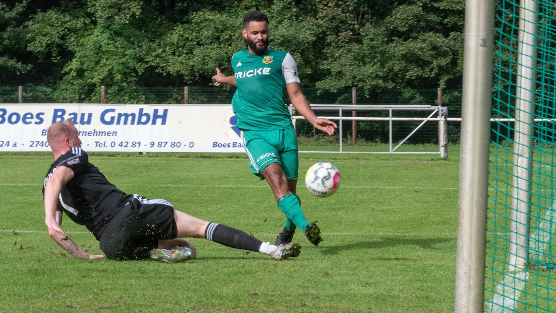 Ein Mann in einem grünen Trikot schießt einen Fußball auf ein Tor und ein Mann im schwarzen Trikot versucht das zu verhindern.