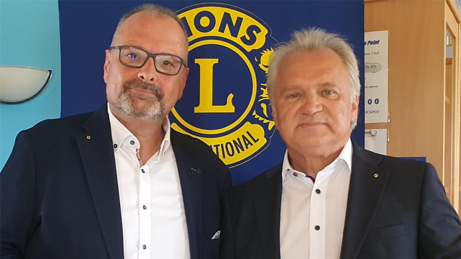 Zwei Männer in weißen Hemden und dunklen Sakkos stehen nebeneinander vor dem Logo von Lions International