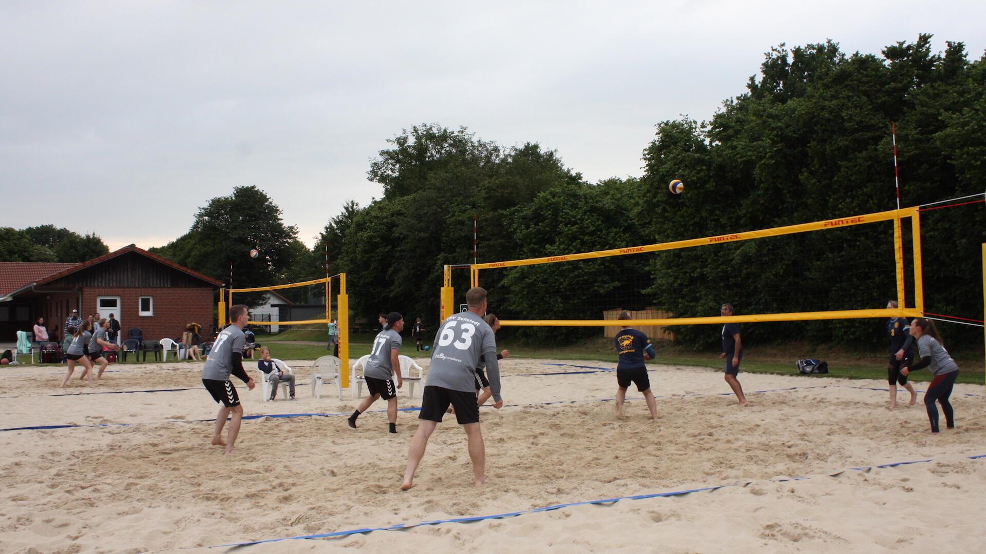 Eine Gruppe Sportler spielt auf zwei nebeneinander liegenden Sandplätzen Beachvolleyball