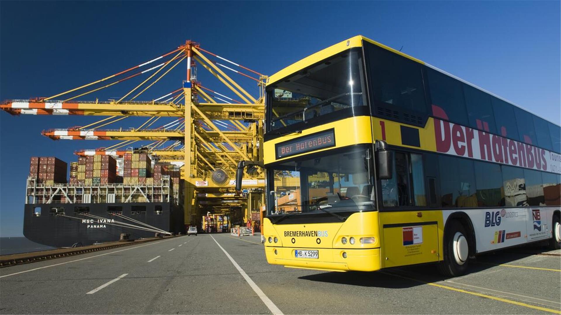 Ein gelber Doppeldecker Bus steht vor einem Containerschiff im Hafen. Die Sonne scheint.