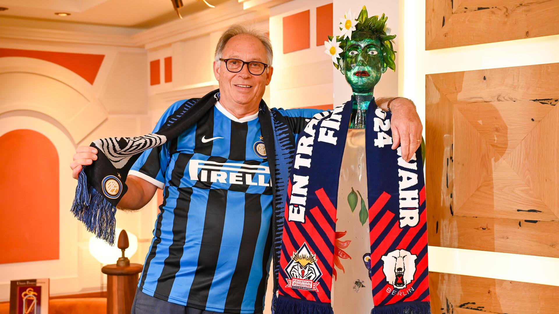 Ein älterer Mann, der ein schwarz-blaues Fußballtrikot anhat und einen blau-roten Eishockeyschal in der Hand hat. 