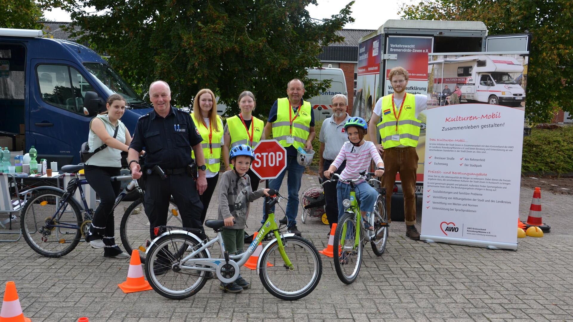 Eine Gruppe von sechs stehenden Menschen und drei Kindern und Jugendlichen mit dem Fahrrad posieren vor einem Stoppschild.