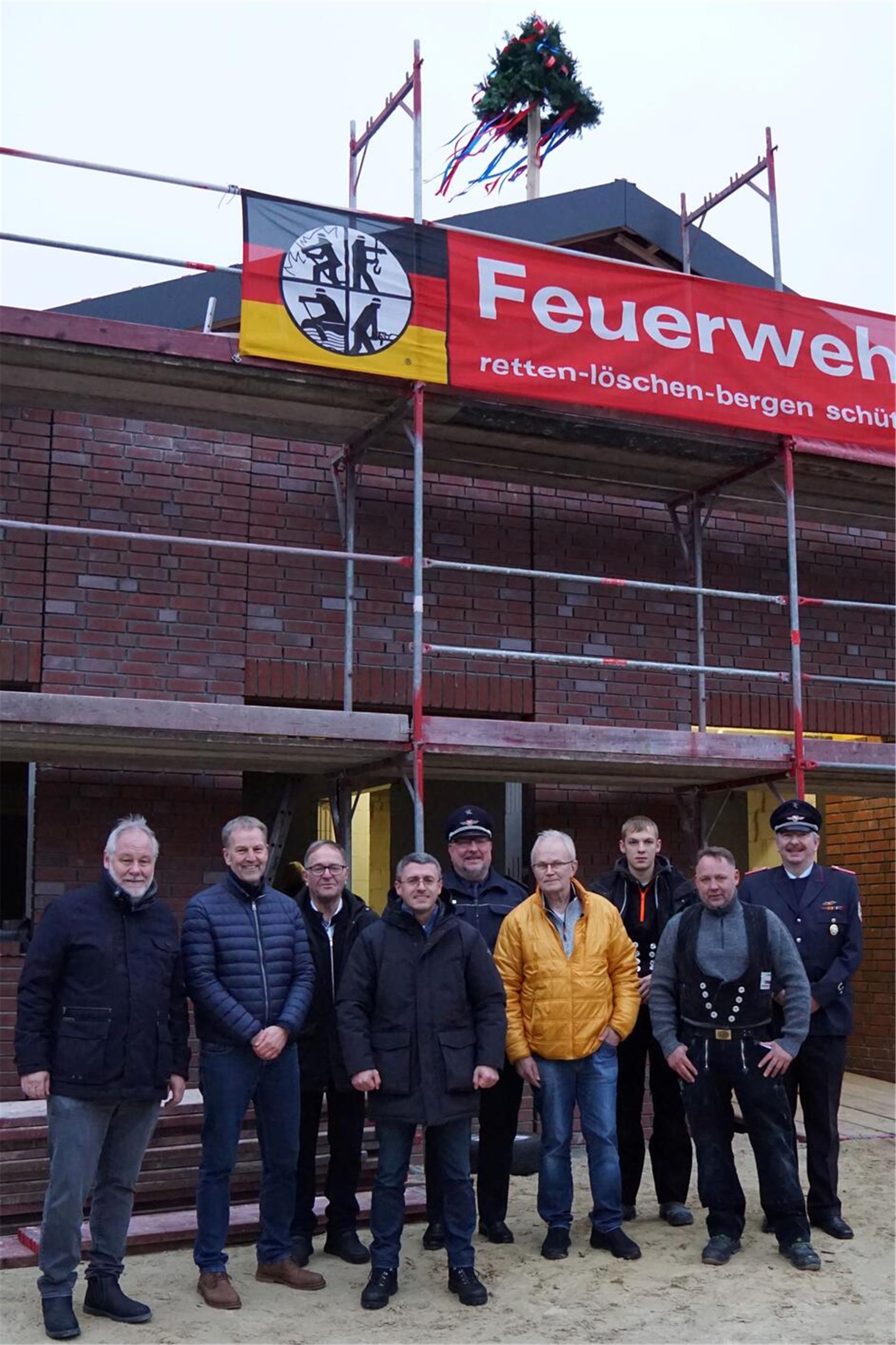 Eine Gruppe von Männern steht vor einem im Bau befindlichen Gebäude, das mit einem Gerüst und einem Banner der Feuerwehr mit der Aufschrift „retten-löschen-bergen-schützen“ versehen ist. Auf dem Dach des Gebäudes ist ein geschmückter Richtkranz angebracht.