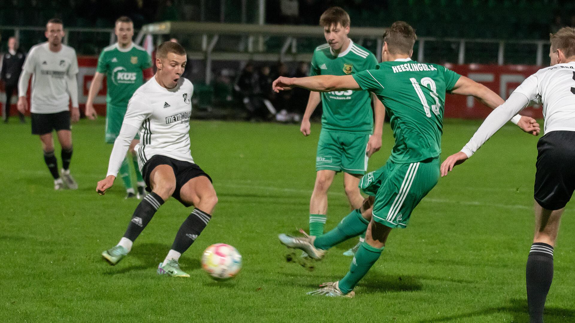 Zwei Männer, einer in einem weißen Trikot und einer in einem grünen Trikot, spielen sich ein Ball zu und mehrere Männer stehen drumherum.