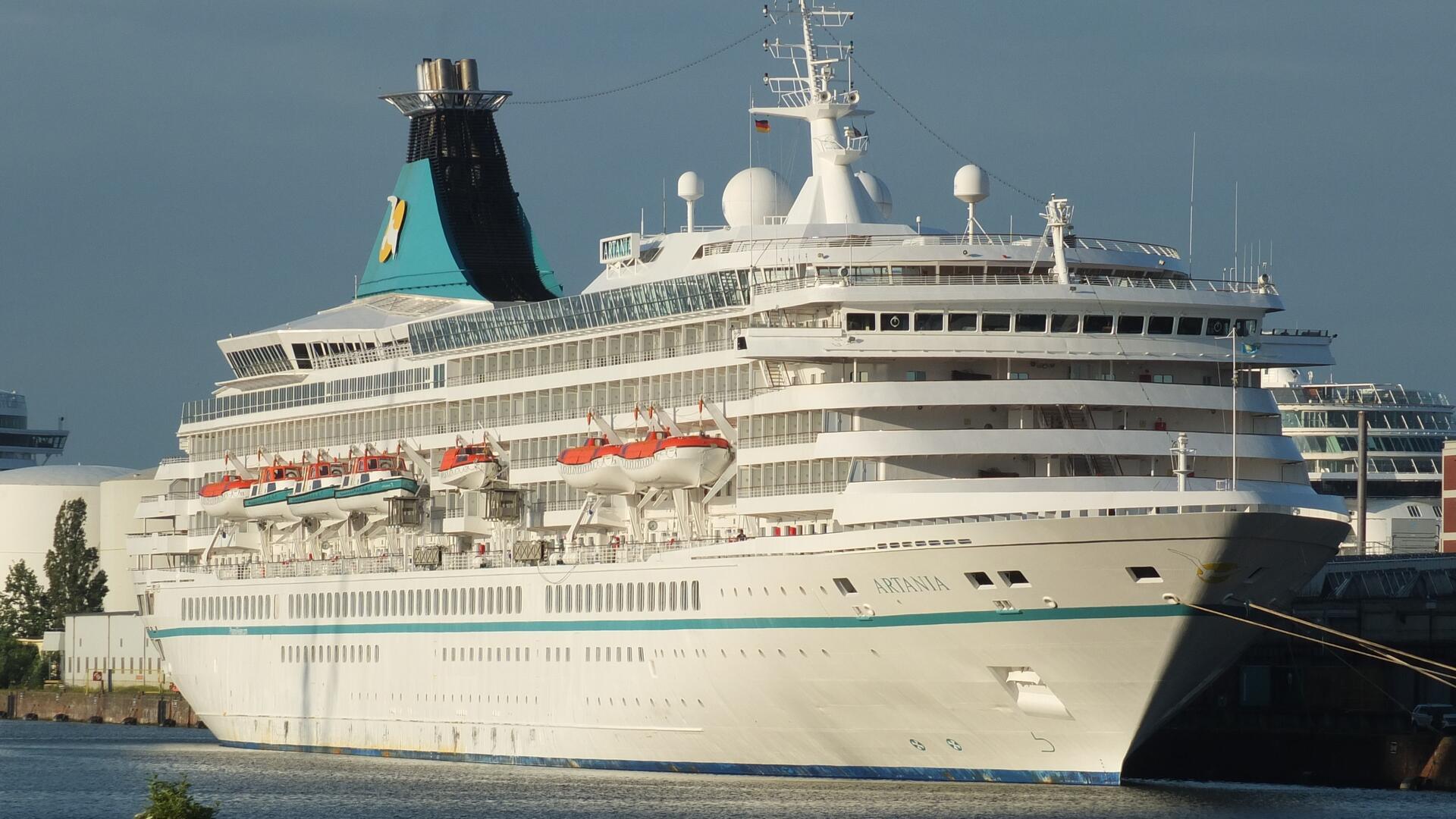 Das Kreuzfahrtschiff „Artania“ liegt im Hafen. Das weiße Schiff mit türkisfarbenen Details verfügt über mehrere Decks, auf denen Rettungsboote zu sehen sind. Der markante Schornstein trägt das Logo von Phoenix Reisen.