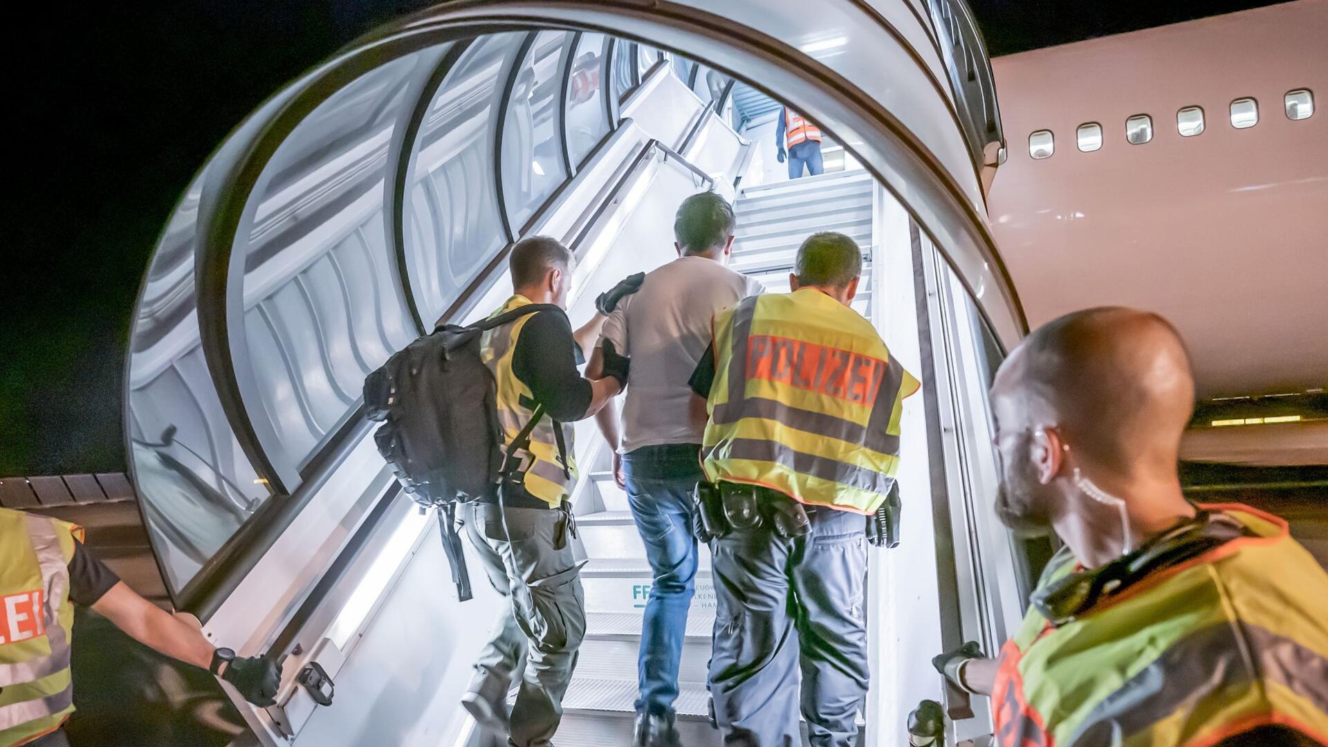 Polizisten führen einen Mann die Gangway zum Flugzeug hinauf.