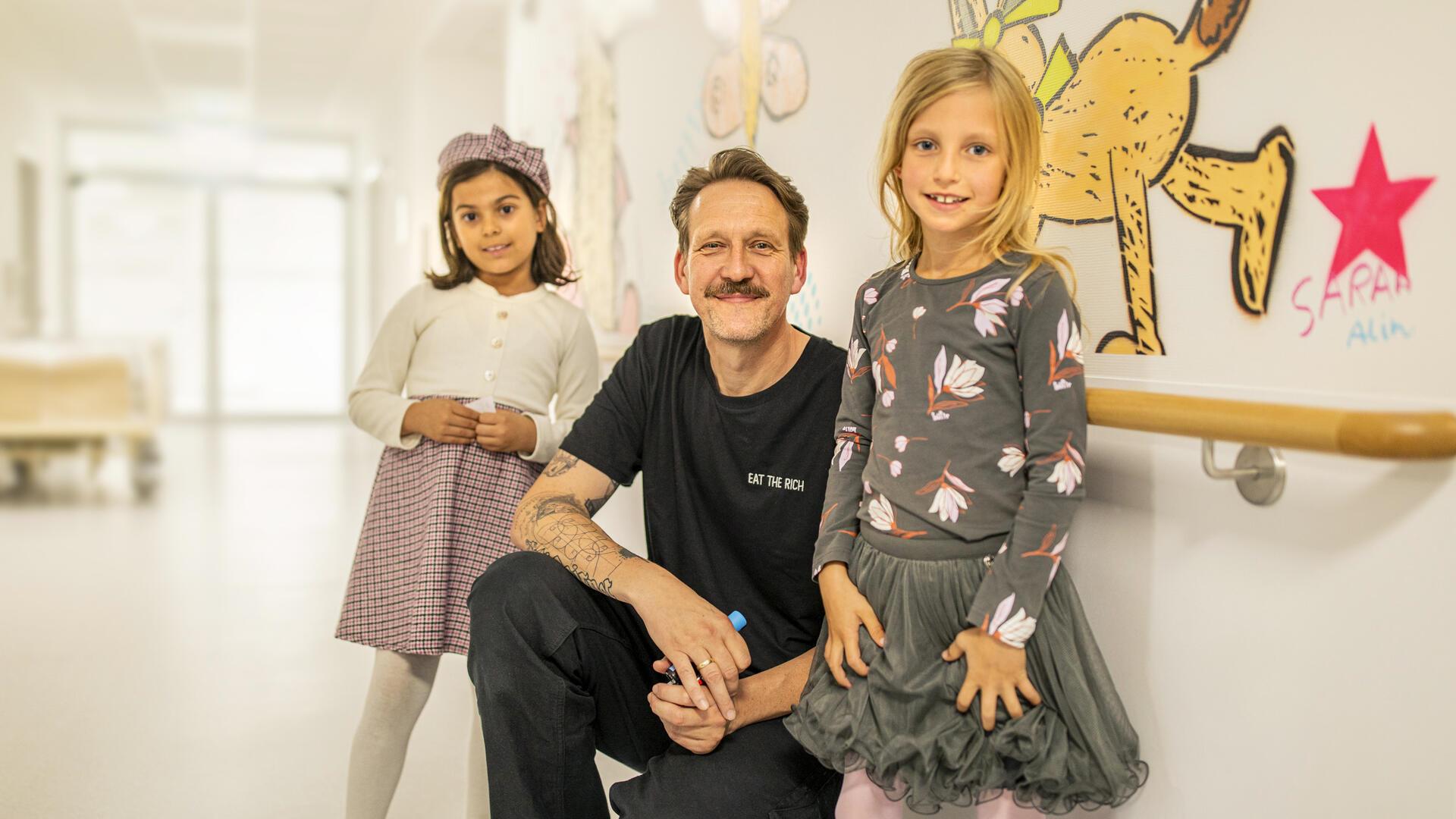 Nils Klempow von dem Künstlerduo Super 8 hat die Zeichnung der Kinder vergrößert auf die Wände der neuen Kinderstation Bremerhaven-Reinkenheide übertragen. Hier zu sehen mit den kleinen Künstlerinnen Alin (links) und Ida (rechts).