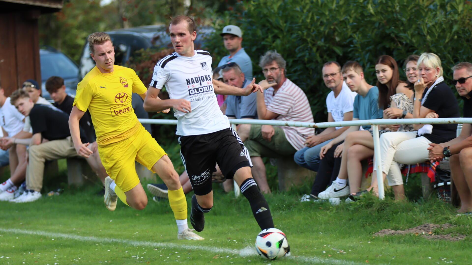 Zwei Männer laufen hinter einem Ball her und Menschen am dahinter guckem zu.