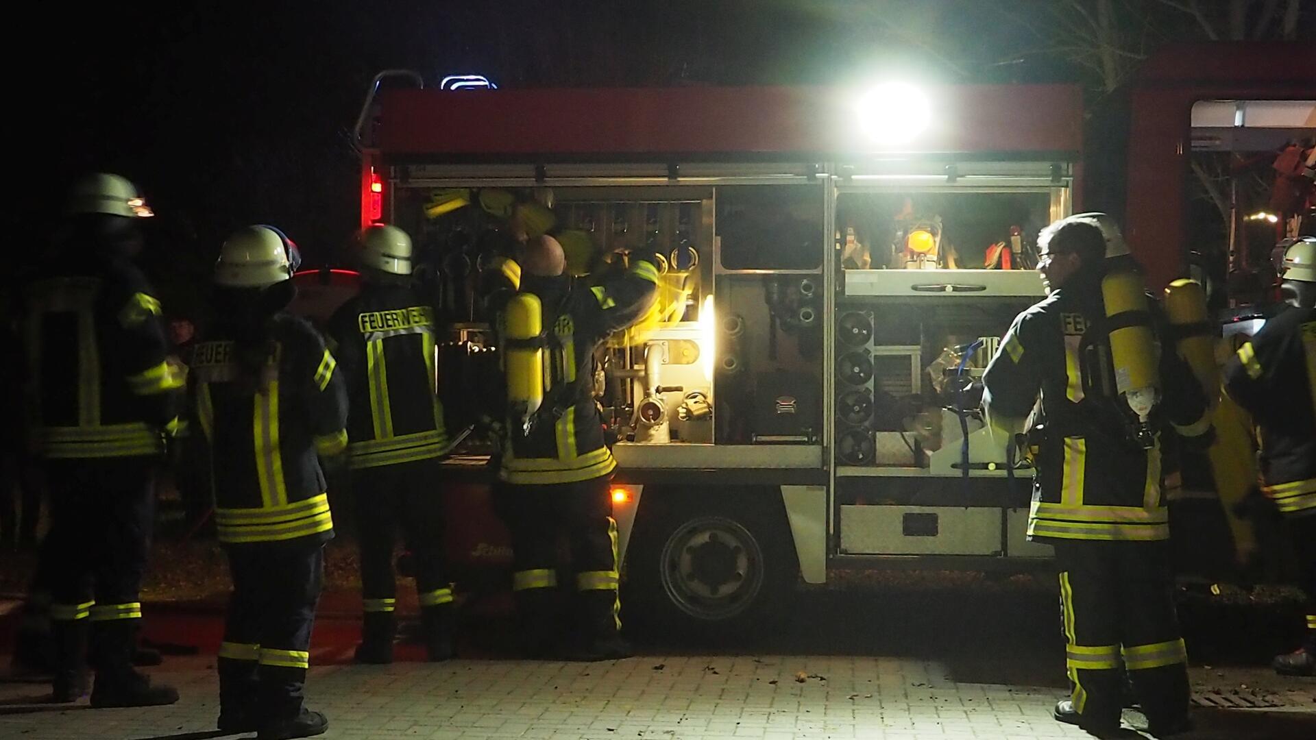 Mehrere Feuerwehrleute in voller Einsatzkleidung stehen neben einem Feuerwehrfahrzeug, dessen hinteres Fach geöffnet ist. Das Fahrzeug ist beleuchtet, und die Feuerwehrleute scheinen sich auf ihren Einsatz vorzubereiten, indem sie Ausrüstung entnehmen.