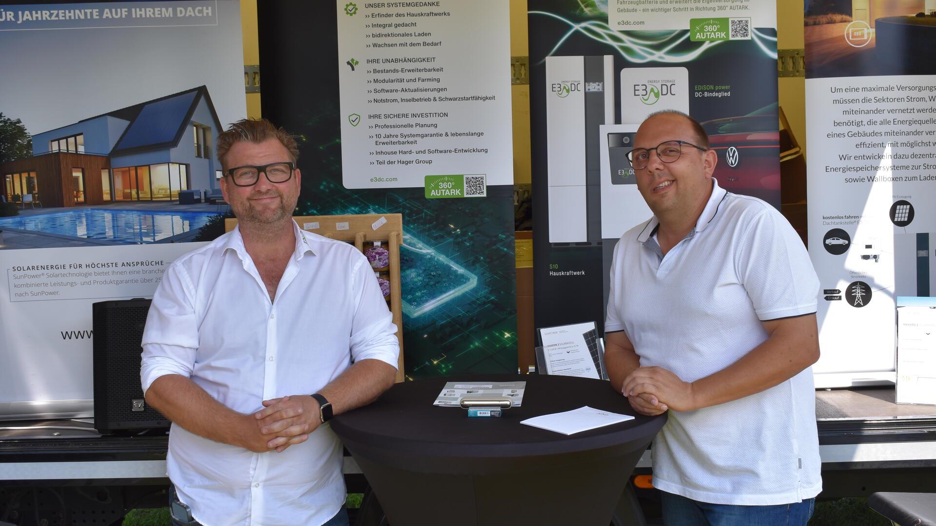 Zwei Männer in weißem Hemd und weißem Poloshirt an einem Stehtisch vor einer Werbewand.