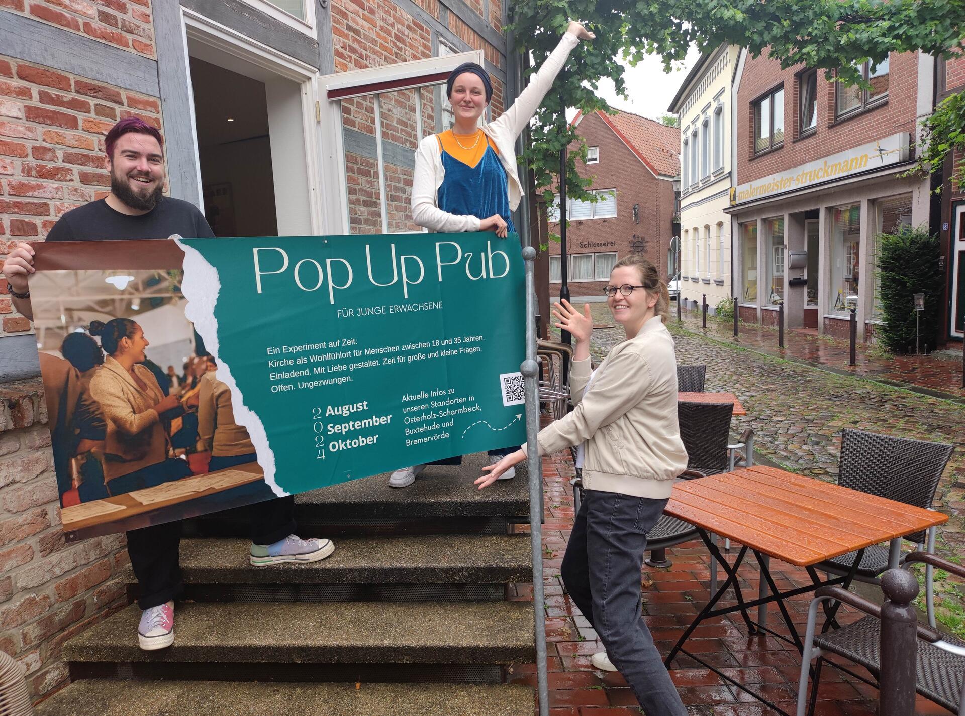 Drei junge Menschen halten ein Plakat in der Hand, auf dem "Pop Up Pub" steht.