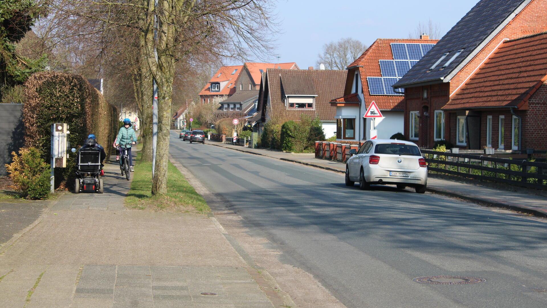 In der Altluneberger Straße parken Autos am Straßenrand, während sich auf dem Gehweg ein Rollstuhlfahrer und ein Fahrradfahrer begegnen.