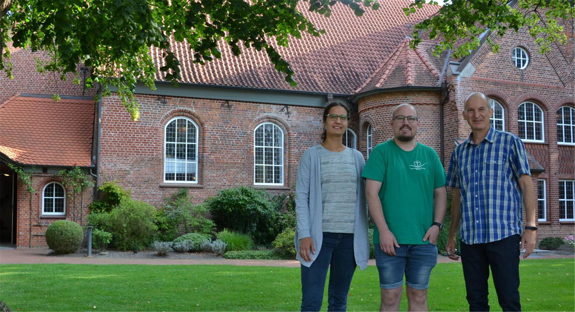 Eine Frau und zwei Männer blicken in die Kamera, hinter ihnen ist ein Kirchengebäude erkennbar.