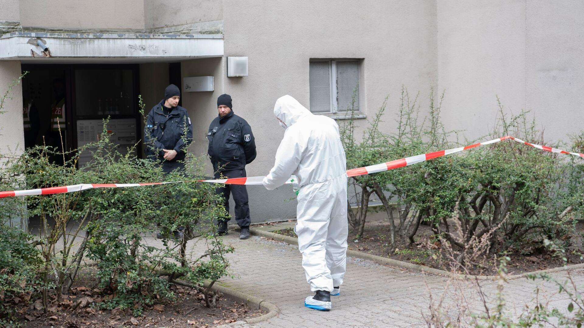 Polizisten stehen am Eingang eines Mehrfamilienhauses in Stadtteil Kreuzberg, während ein Polizist in Schutzanzug an einem Absperrband steht.