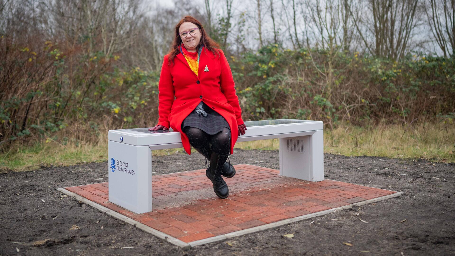 Eine Frau in einem leuchtend roten Mantel sitzt auf einer modernen weißen Sitzbank mit Solarflächen, die auf einem kleinen gepflasterten Bereich steht. Die Bank trägt das Logo der "Seestadt Bremerhaven".