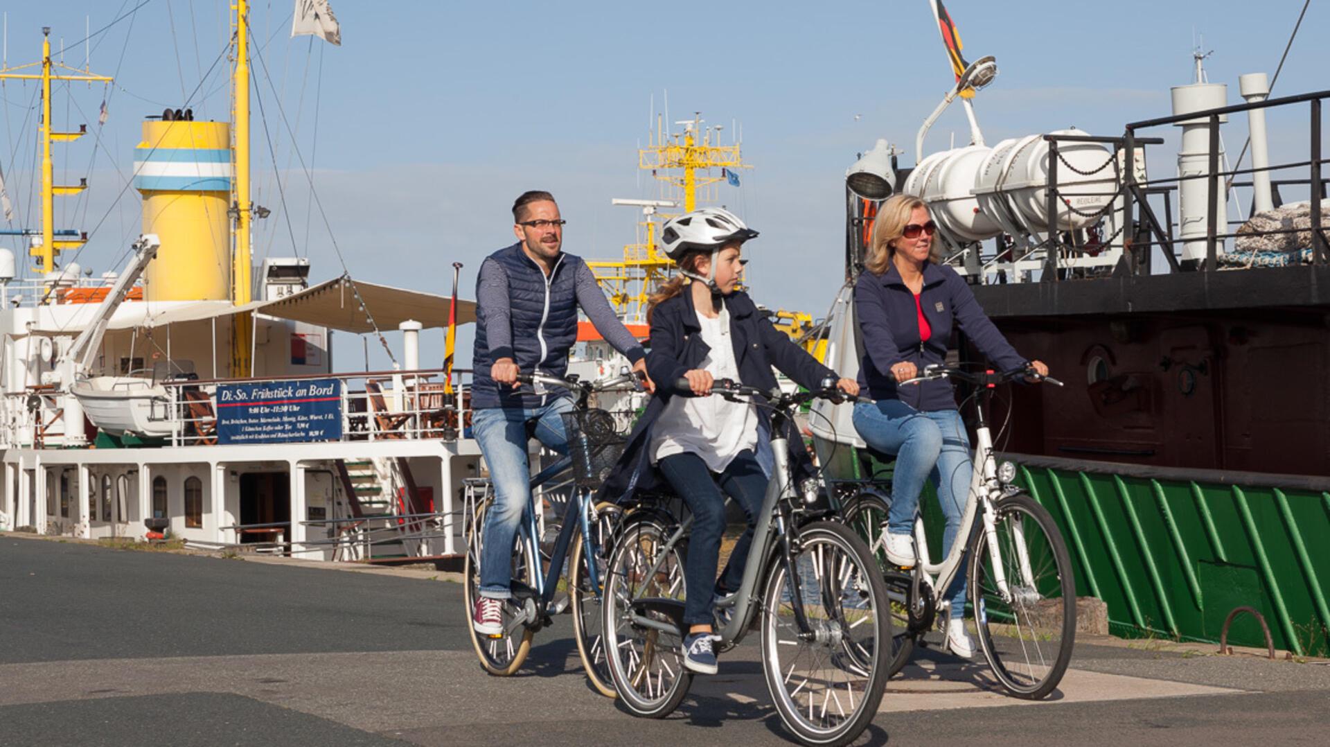 Eine Familie auf dem Fahrrad fährt an Schiffen im Hafen entlang.