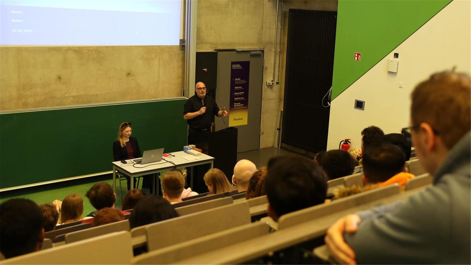 ein Mann an einem Rednerpult wendet sich an Studentinnen und Studenten in einem Hörsaal