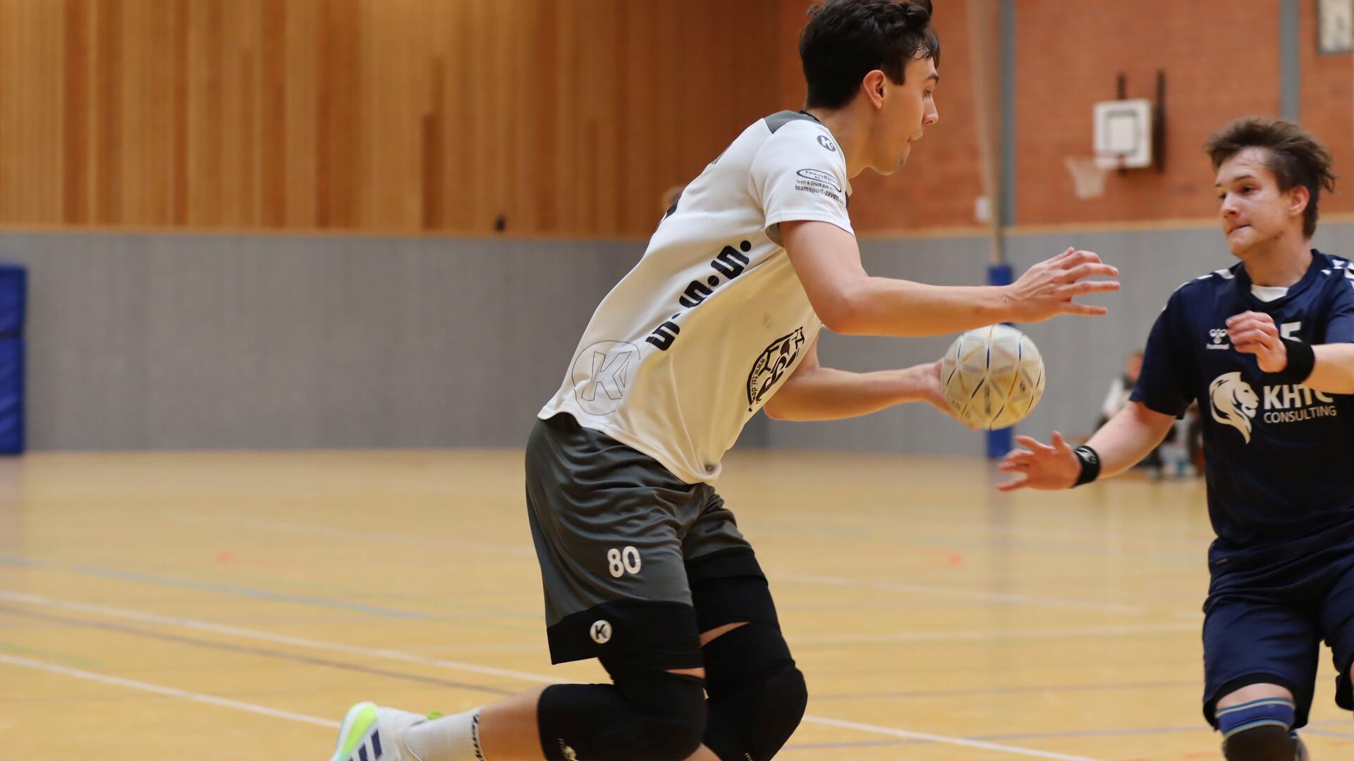 Ein junger Mann in schwarzweißem Trikot hält in einer Sporthalle einen weißen Ball in der Hand, den versucht ein zweiter Mann in blauem Trikot ihm abzunehmen.