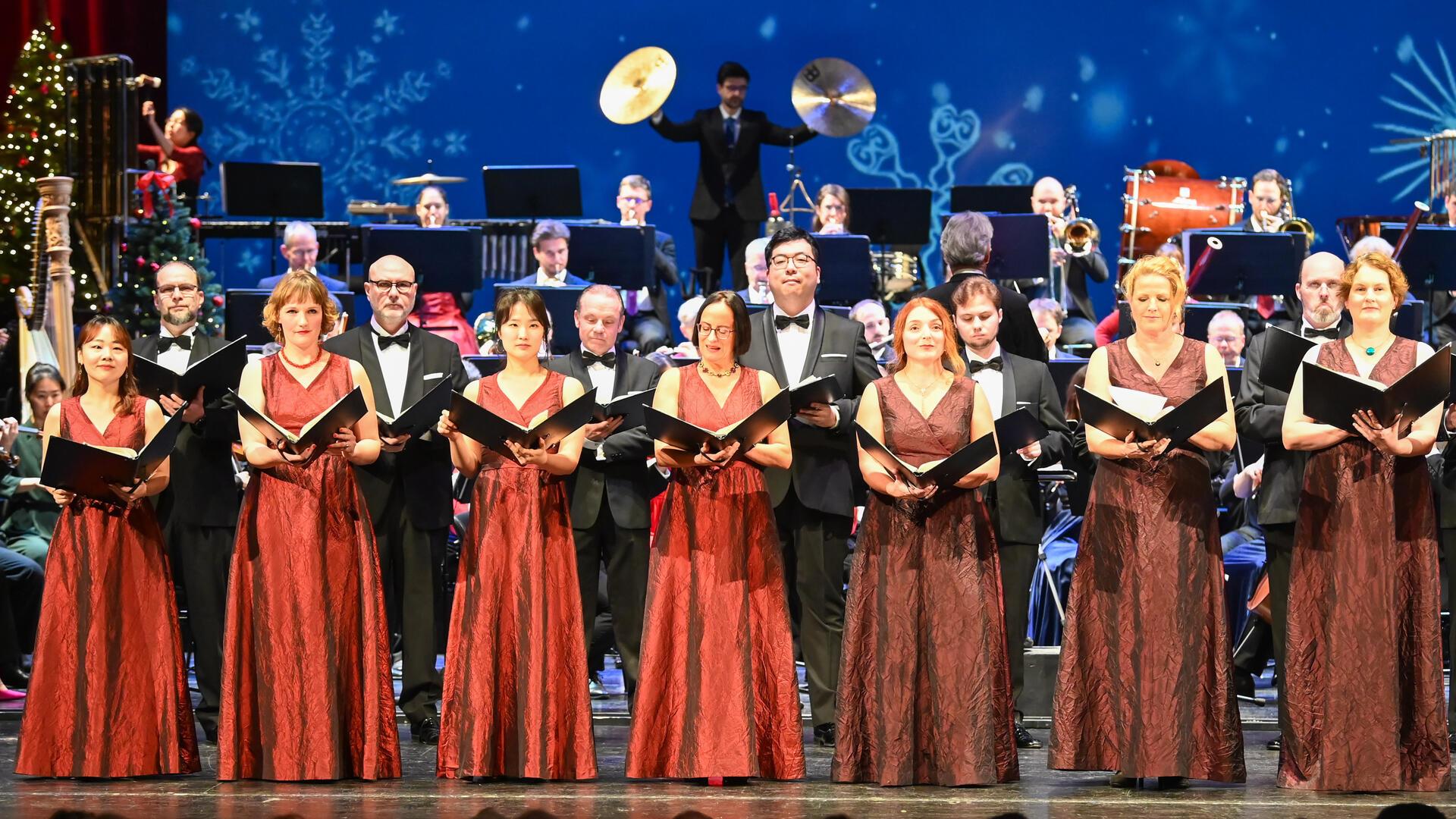 Ein Chor, bestehend aus Männern und Frauen, trägt elegante Abendkleidung. Die Frauen tragen rote, bodenlange Kleider, die Männer schwarze Anzüge mit Fliege. Sie halten Notenblätter und singen vor einem großen Orchester im Hintergrund. Der Bühnenhintergrund zeigt winterliche Motive, darunter Schneeflocken und festliche Dekorationen.