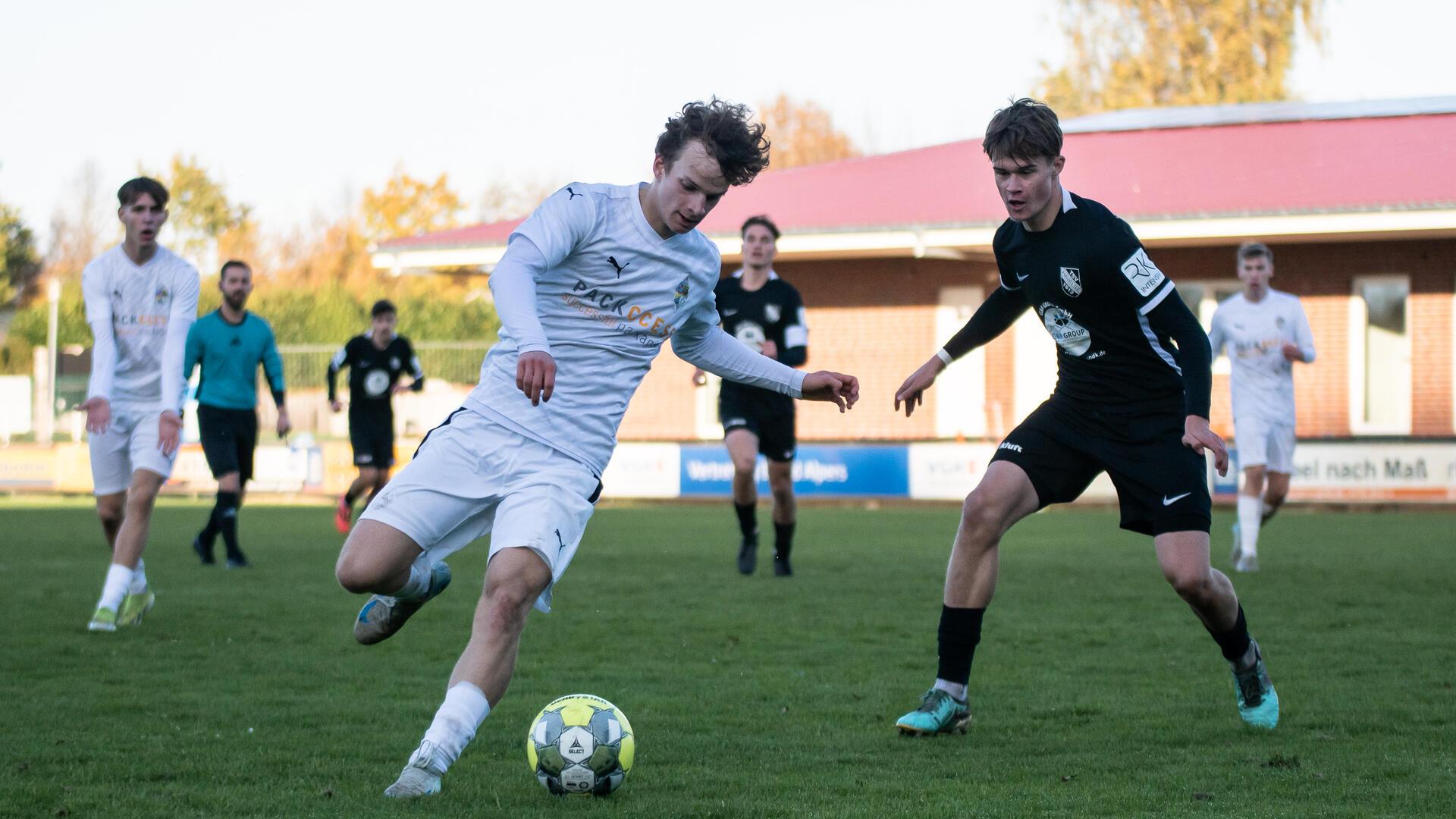 Ein Mann in einem weißen Trikot hat einen Ball ein den Füßen und ein Mann im schwarzen Trikot steht hinter ihm.