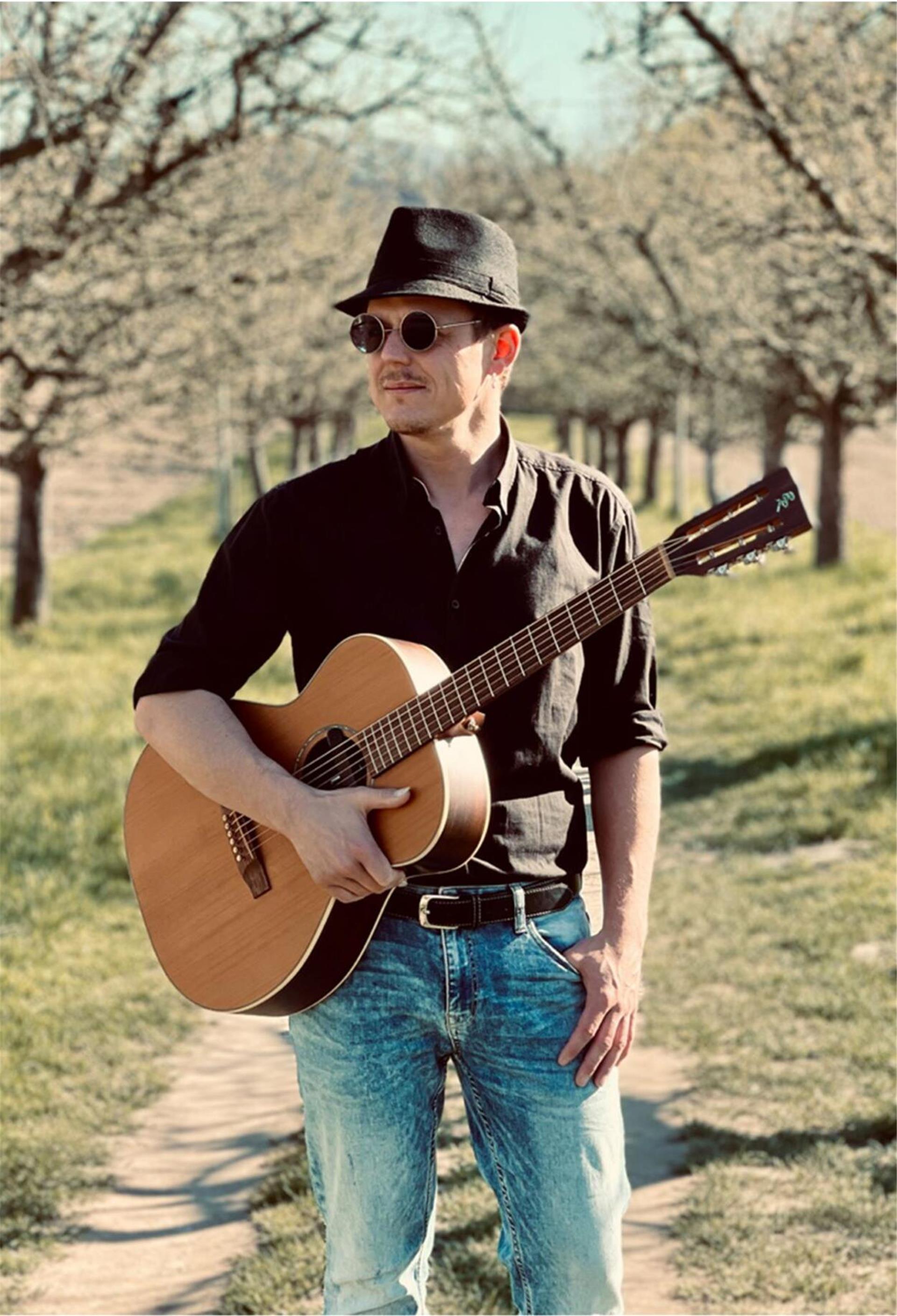 Mann mit Gitarre in der Hand, Sonnebrille und Hut steht in der Landschaft 