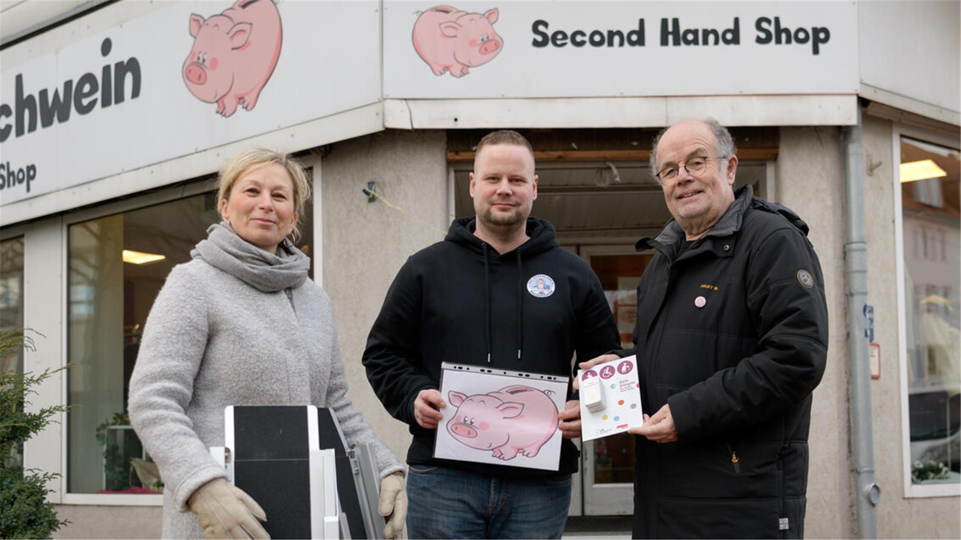 Eine Frau und zwei Männer stehen vor dem Eingang eines Geschäfts. Sie halten verschiedene Dinge in Händen, eine Klingel, ein Bild eines Sparschweins sowie eine mobile Rampe.