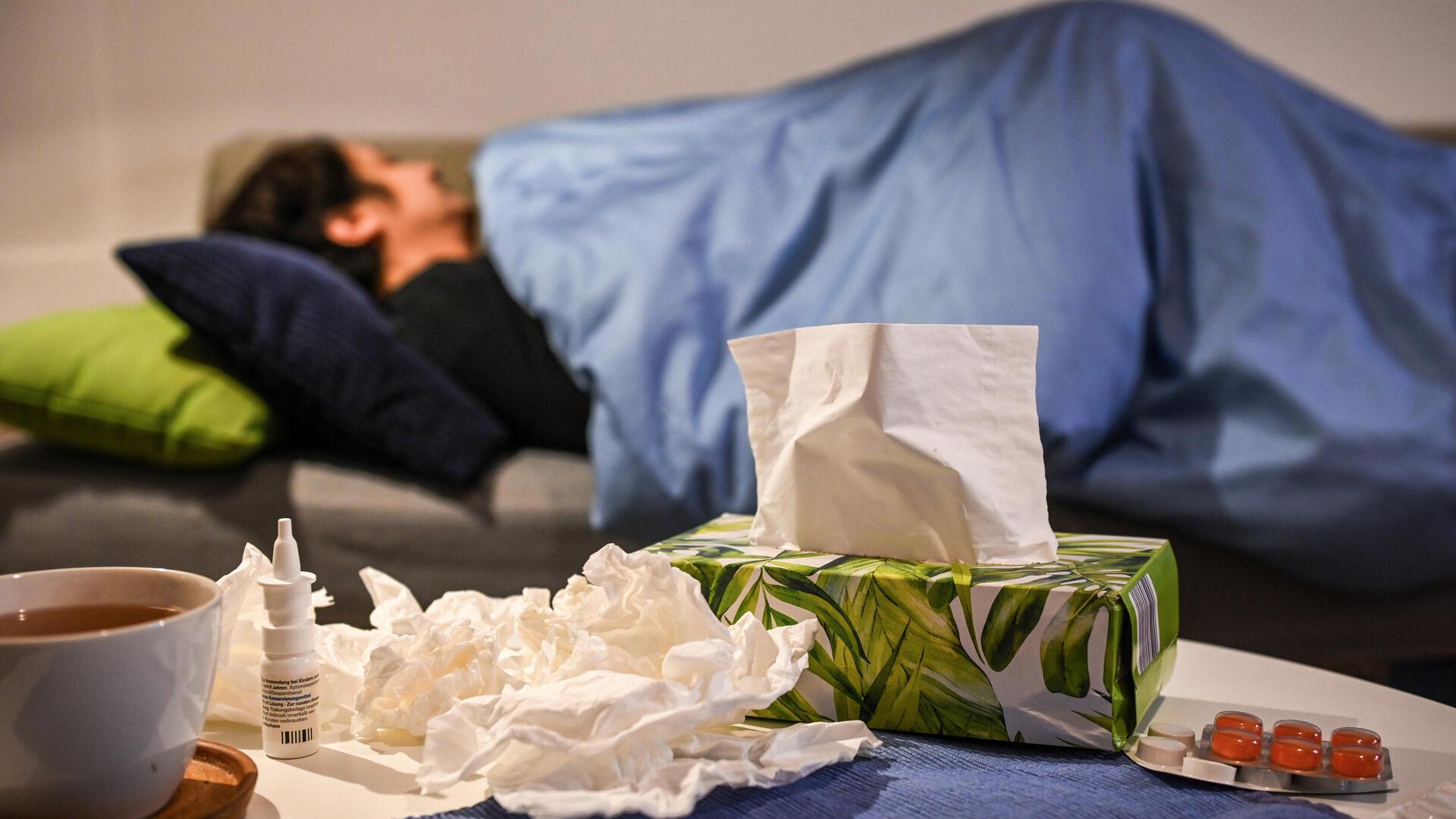 Eine erkrankte Frau liegt unter einer Decke auf dem Sofa. Auf einem Tisch liegen Medikamente gegen Schnupfen und Erkältung - Nasenspray und Halstabletten - außerdem eine Tasse Tee und Taschentücher.