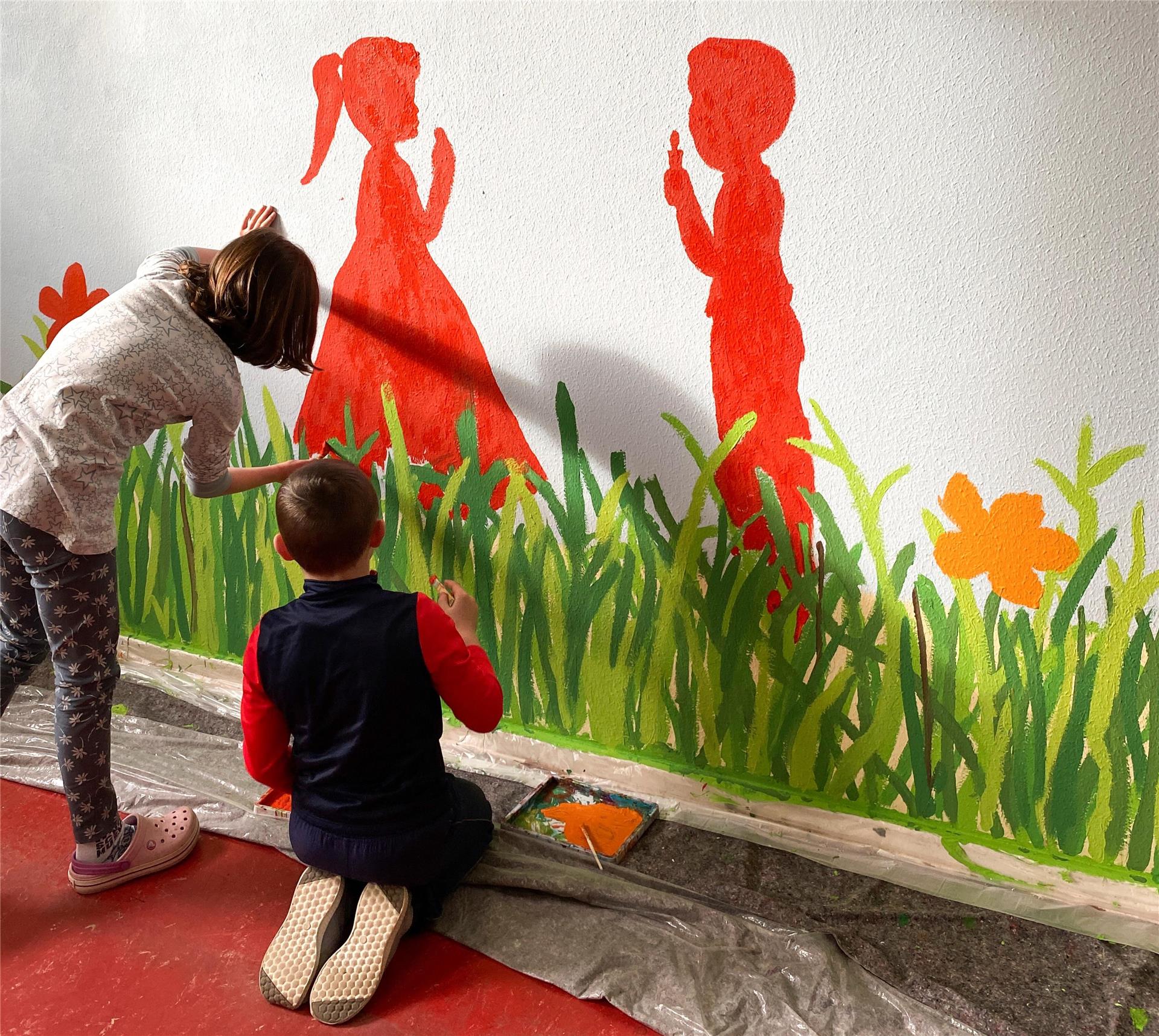 Ein Mädchen und ein Junge malen mit Pinseln eine grüne Wiese an die Wand 