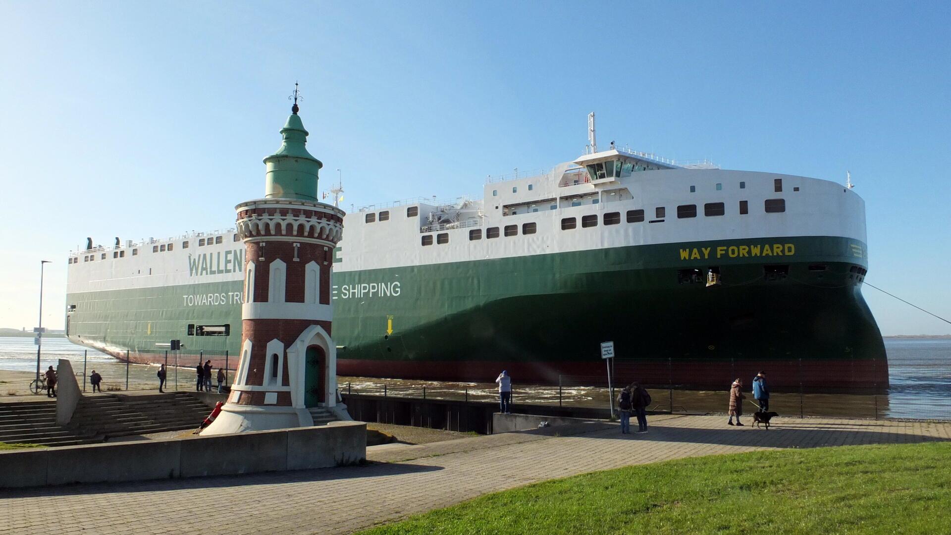 Ein großes grünes Frachtschiff mit der Aufschrift „WAY FORWARD“ und „WALLENIUS SHIPPING“ fährt an einer Promenade entlang, während einige Personen auf dem Weg und am Ufer stehen und das Schiff beobachten. Im Vordergrund ist der Bremerhavener Pingelturm zu sehen.