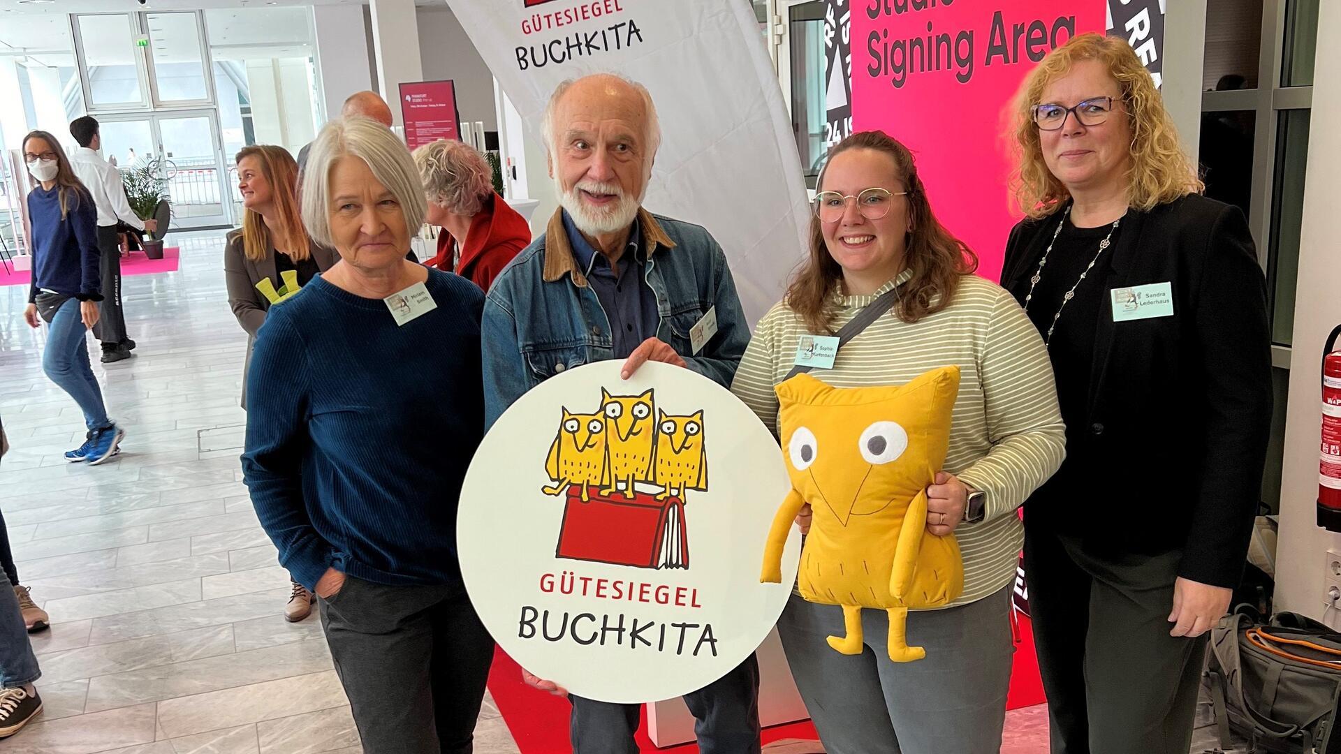 Drei Frauen und ein Mann stehen nebeneinander. Sie halten ein großes Emblem mit der Aufschrift "Gütesiegel Buchkita".