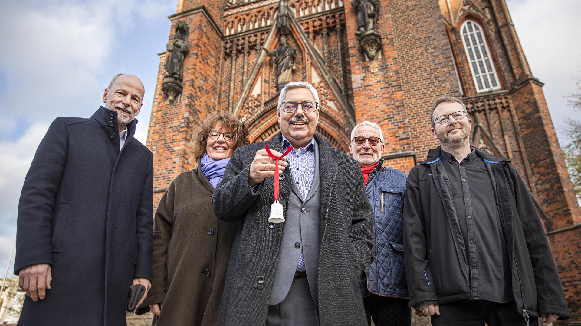 Vier Männer und eine Frau stehen vor der Großen Kirche in Bremerhaven. Ein Mann hält ein kleines weißes Glöckchen in der Hand. 