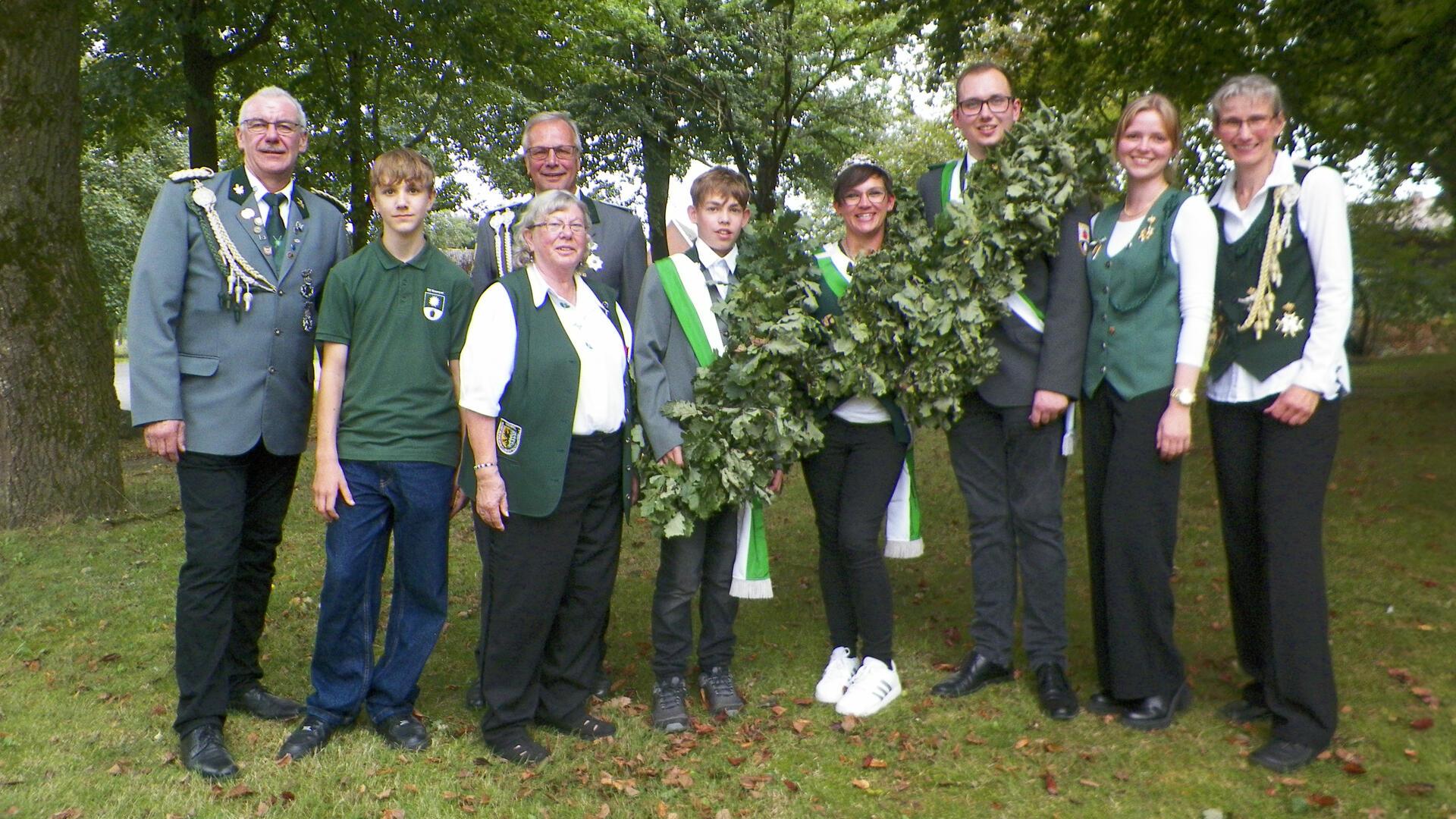 Neun Schützinnen und Schützen in Uniform stehen nebeneinander auf einer Wiese zwischen Bäumen.