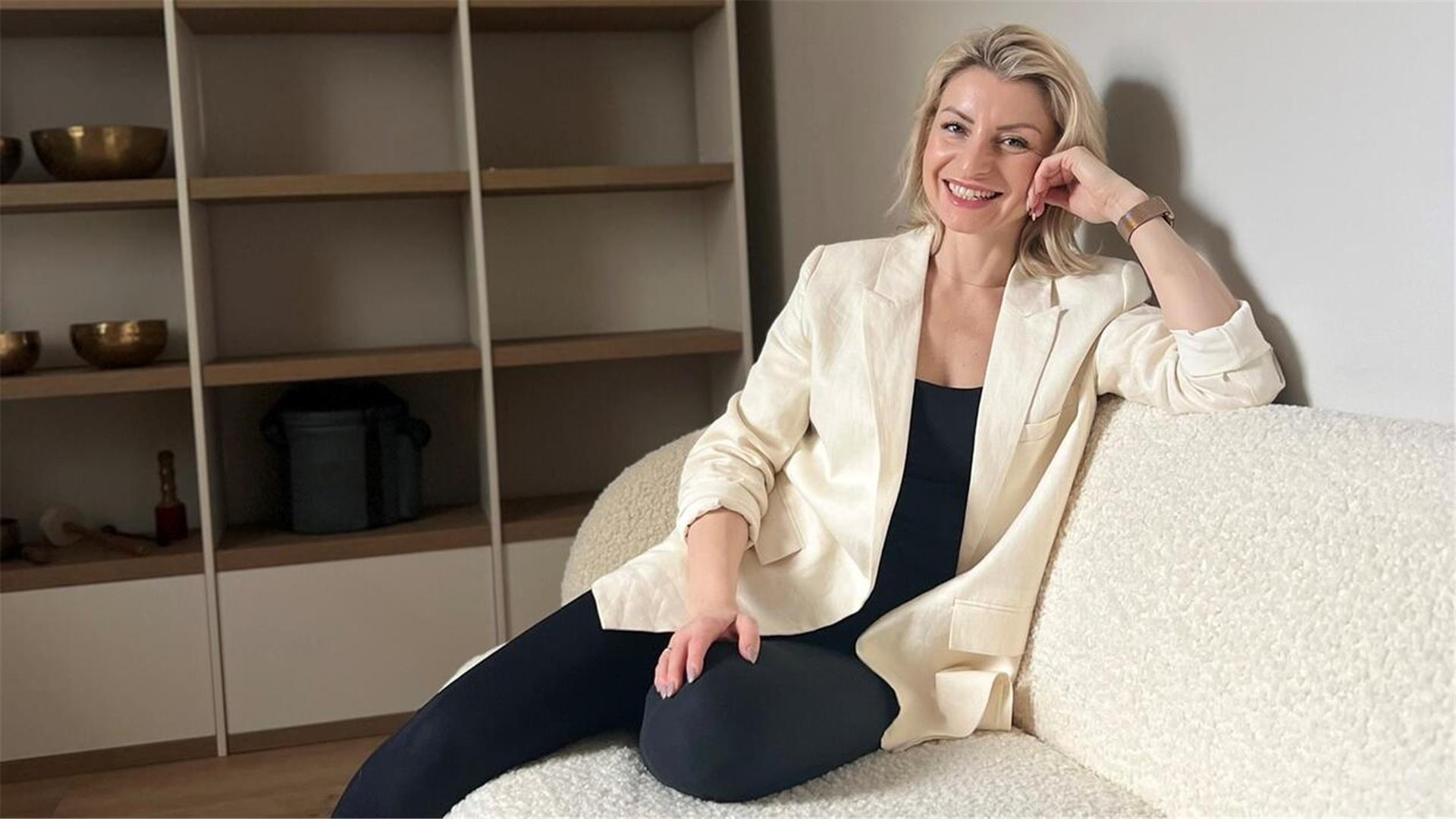 Eine Frau mit blonden Haaren, die einen weißen Blazer und eine dunkle Hose trägt, sitzt auf einer weißen Couch.