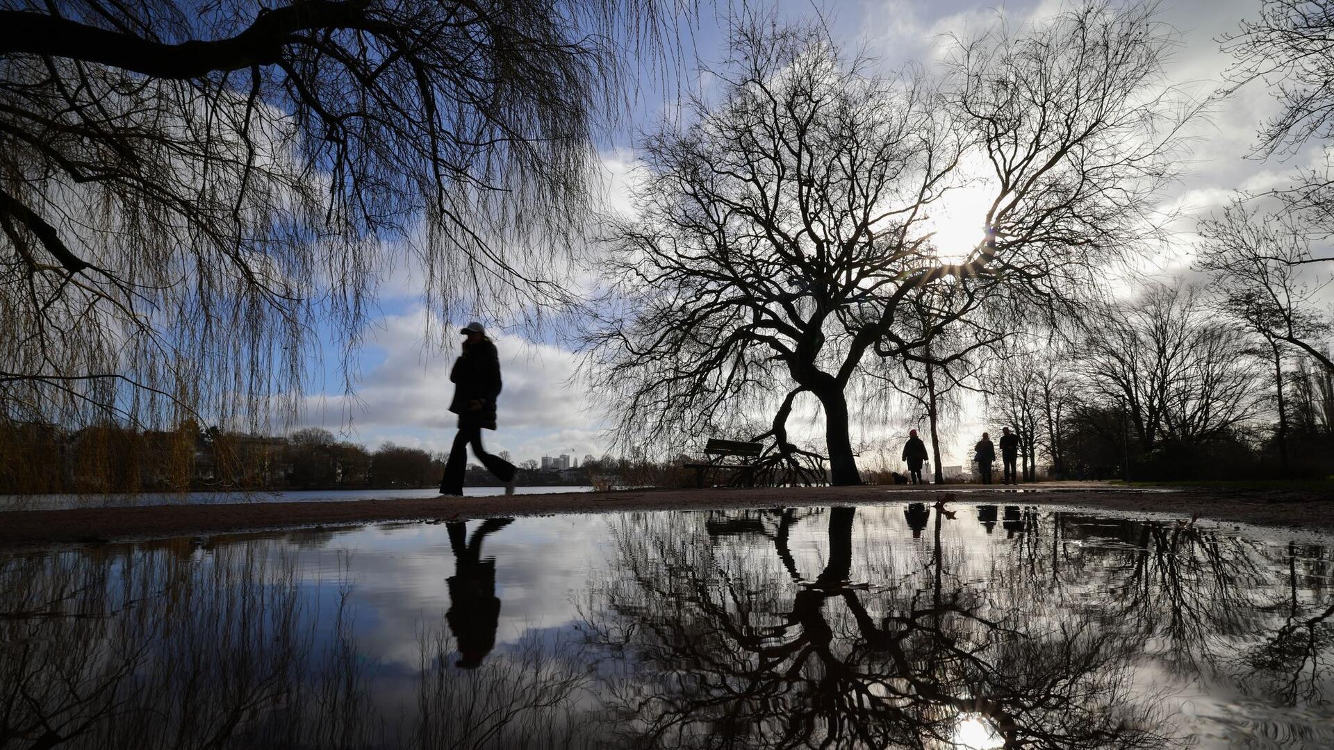 Spiegelung: Spaziergänger genießen den sonnigen Tag an der Alster in Hamburg, ihre Silhouetten und die der kahlen Bäume zeichnen sich in einer Pfütze ab.