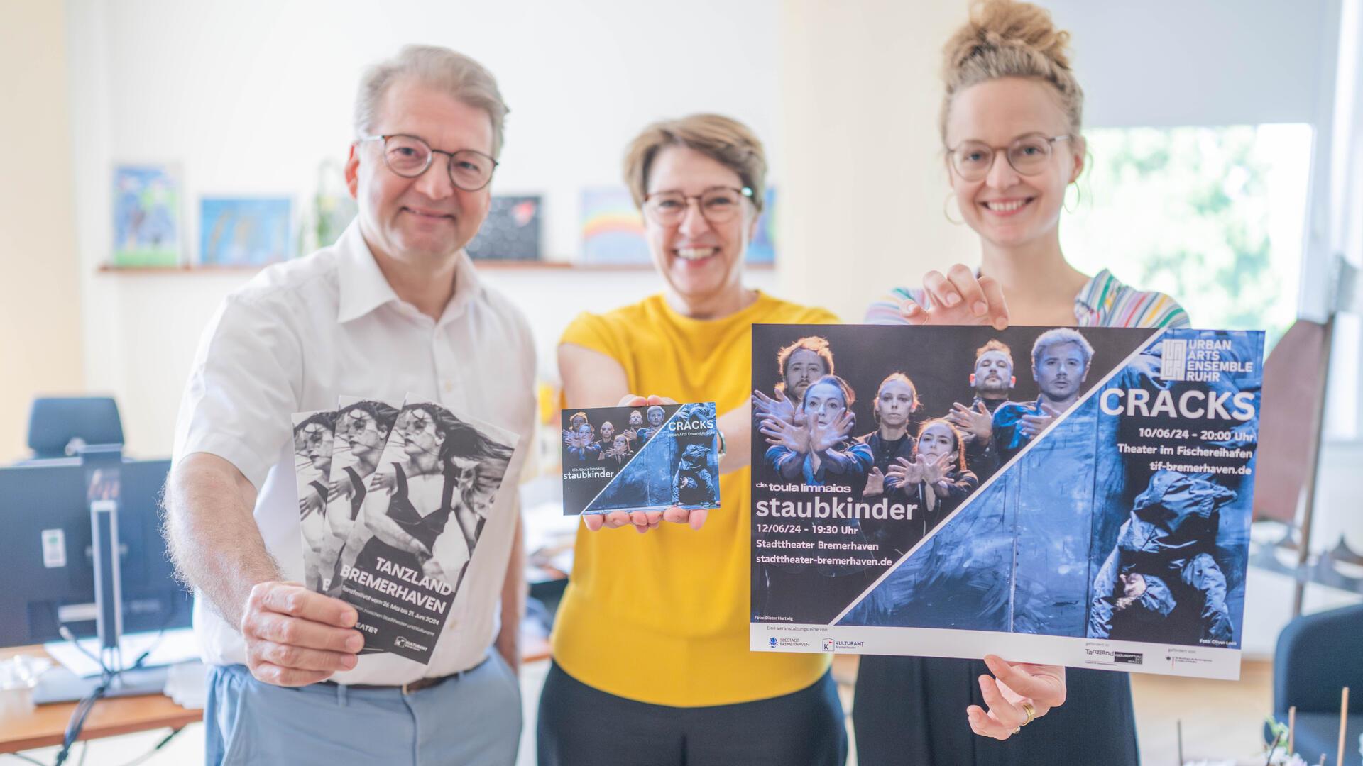 Ein Mann und zwei Frauen stehen lächelnd nebeneinander und halten Flyer und Plakate zum "Tanzland Bremerhaven" in die Kamera