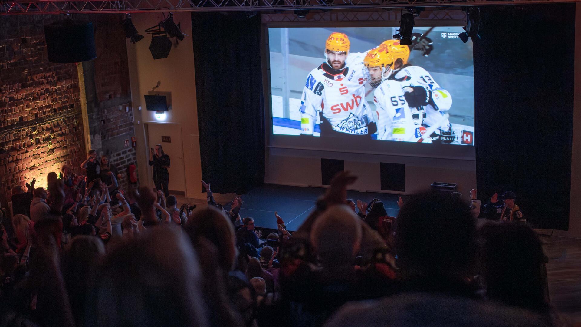 Menge schaut von den Rängen auf einen Bildschirm mit einem Eishockeyspiel. 