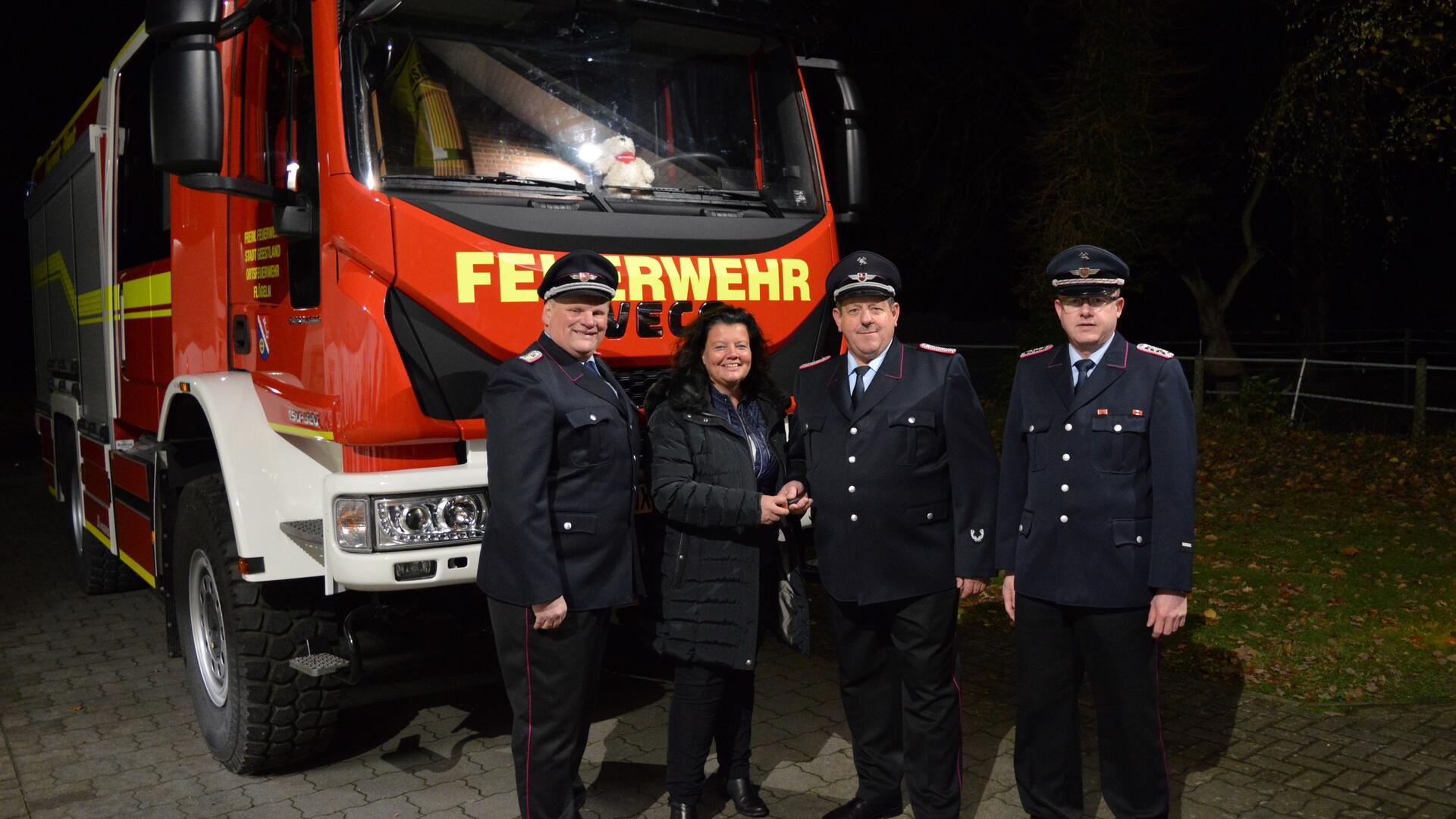 Vier Personen stehen vor einem roten Feuerwehrfahrzeug. Drei Männer in Feuerwehruniformen und eine Frau in dunkler Jacke sind zu sehen. Die Frau übergibt symbolisch einen Schlüssel an einen der Feuerwehrmänner.