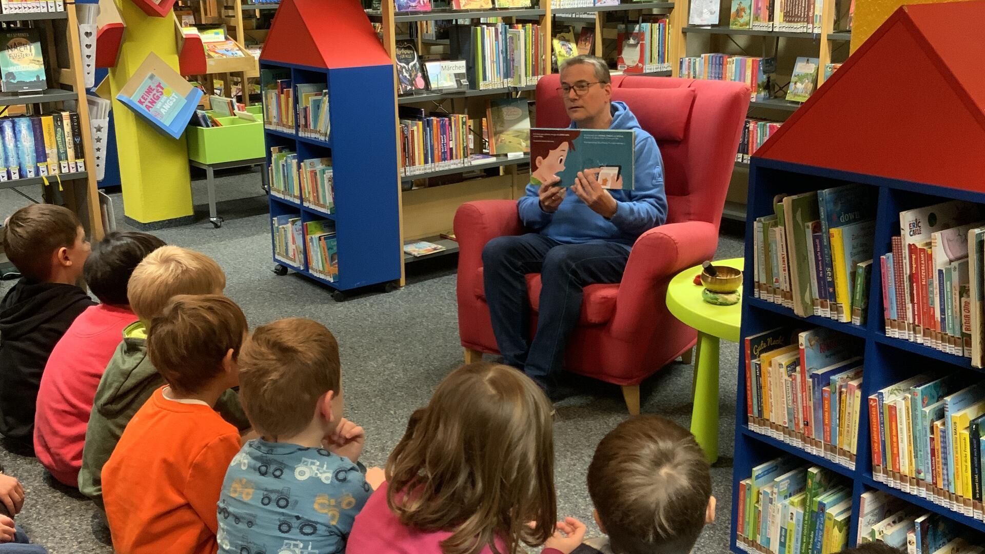 Kinder sitzen in der Bücherei und hören einer Frau zu, die ihnen aus einem Buch etwas vorliest. 