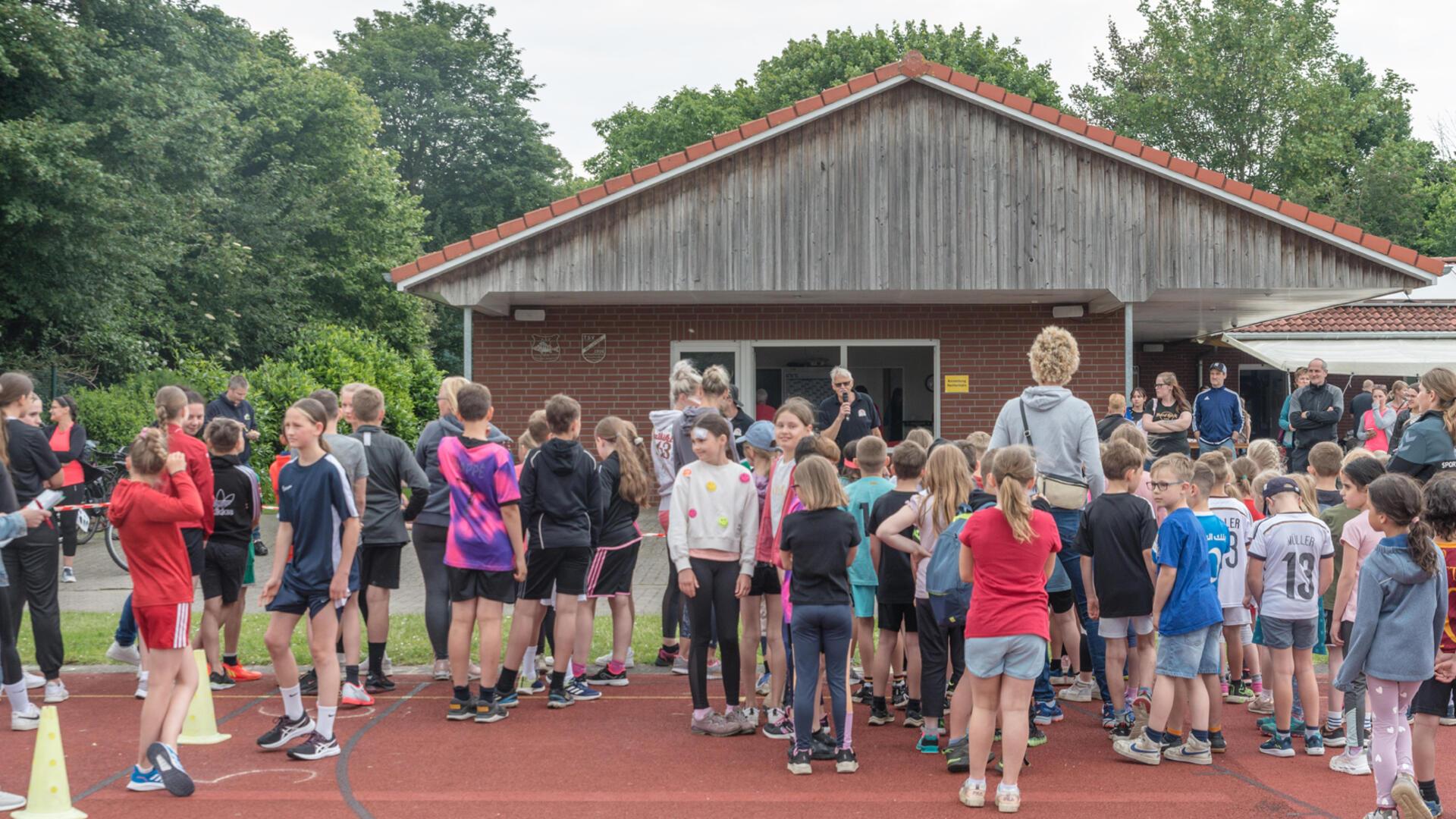 Viele Kinder, Jugendliche und Erwachsene in Sportkleidung stehen auf einer Laufbahn vor einem Gebäude