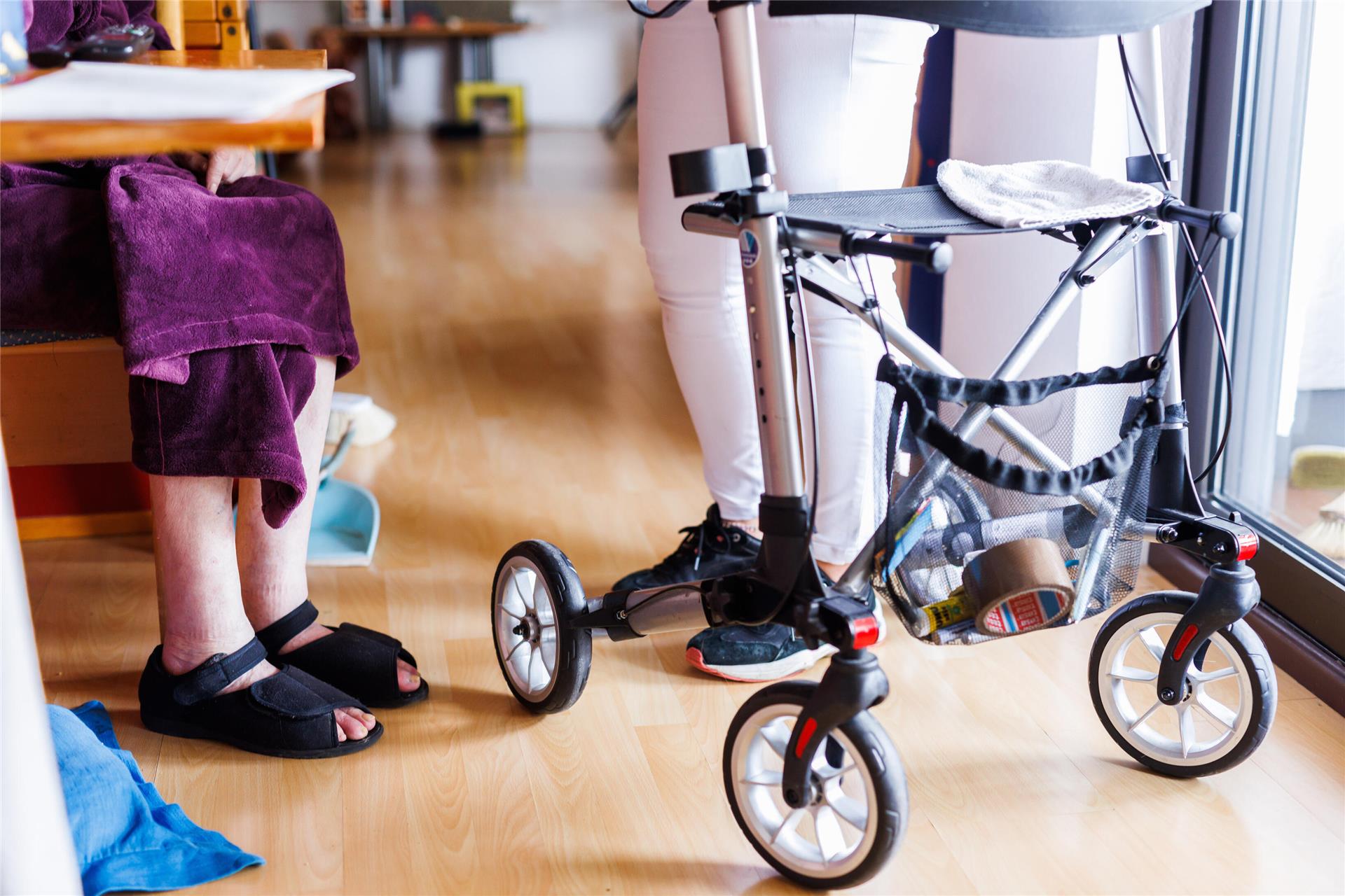 Ein Rollator steht in einem Zimmer. Eine Frau steht, eine weiter sitzt dahinter.